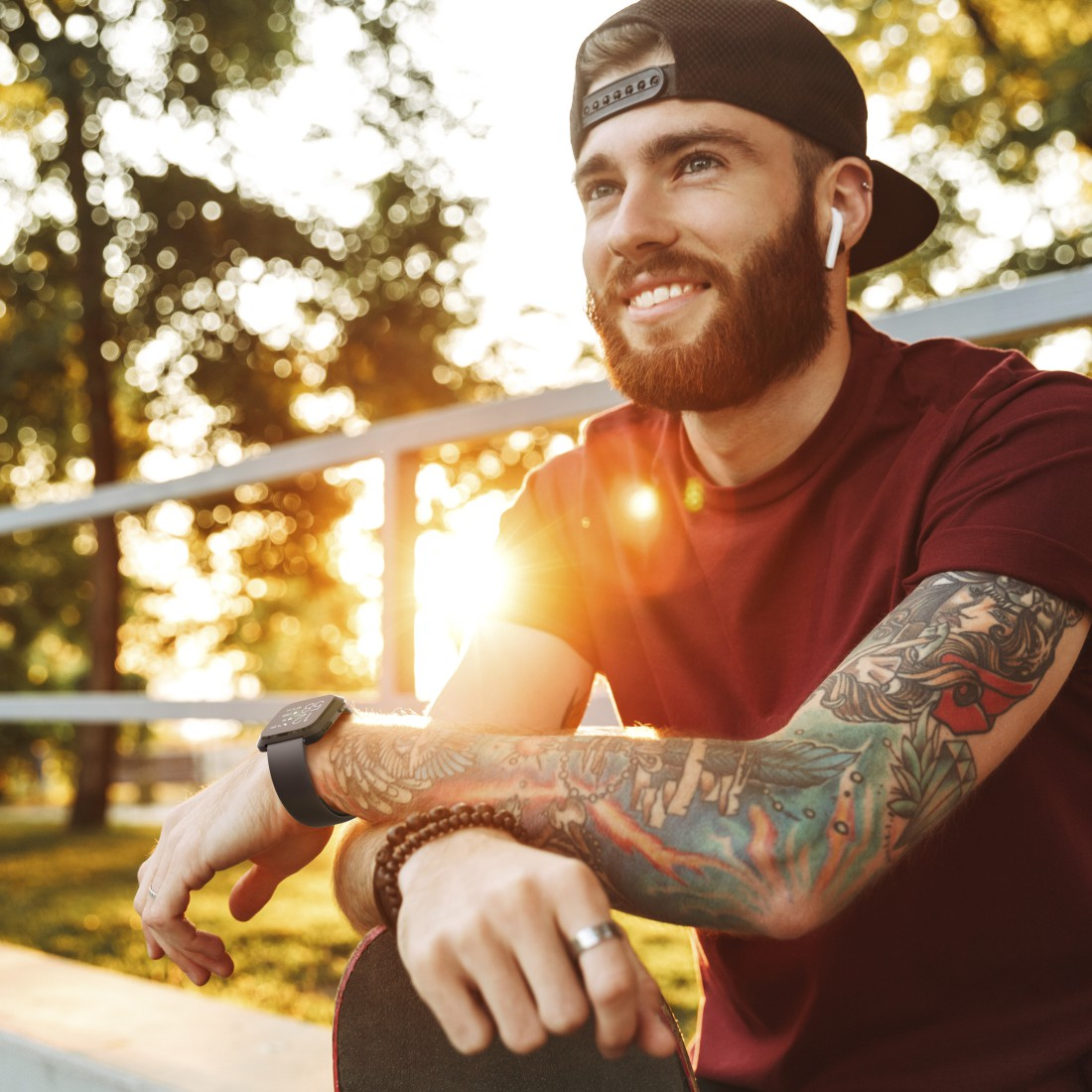 Mann mit Bart und Tattoos, der eine Kappe und Ohrhörer trägt, und mit einem Skateboard in die Sonne schaut.