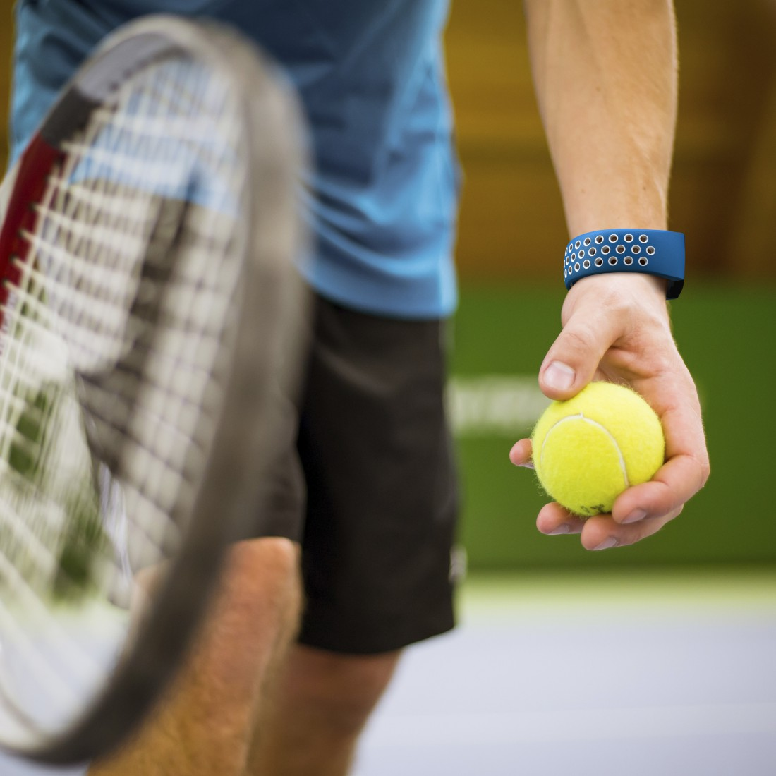 Person hält Tennisball, Tennisschläger und blaues Armband sichtbar.