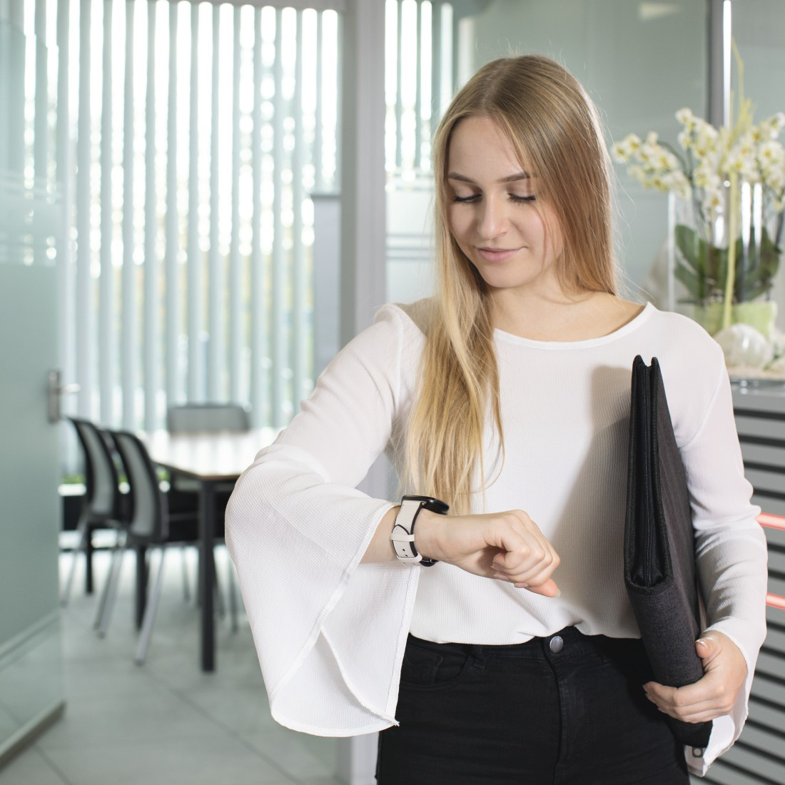 Eine Frau, die in einem Büro auf ihre Smartwatch schaut und eine Aktenmappe hält.