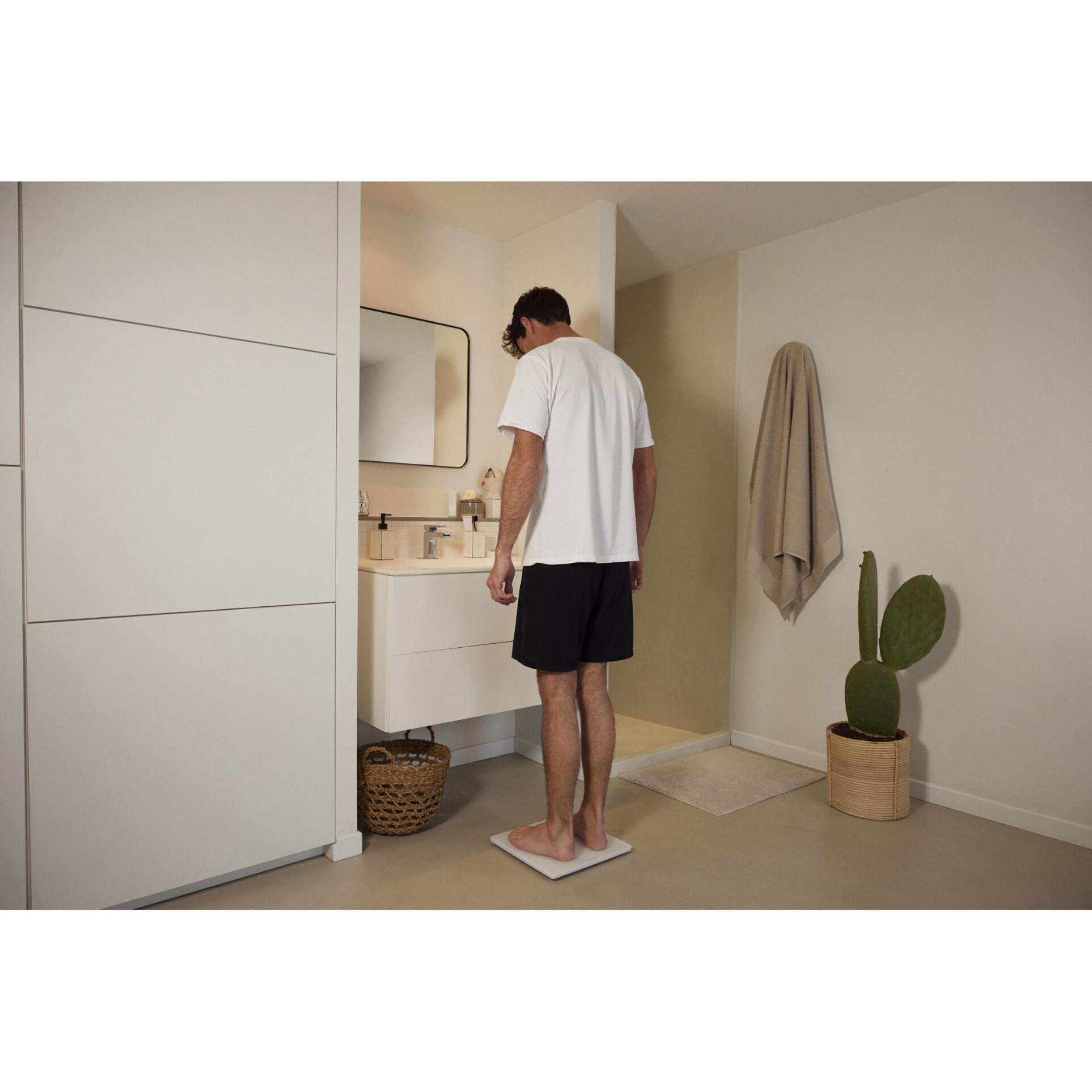 Mann im Badezimmer, trägt Shorts und ein T-Shirt, steht auf einer Waage und schaut nach unten.
