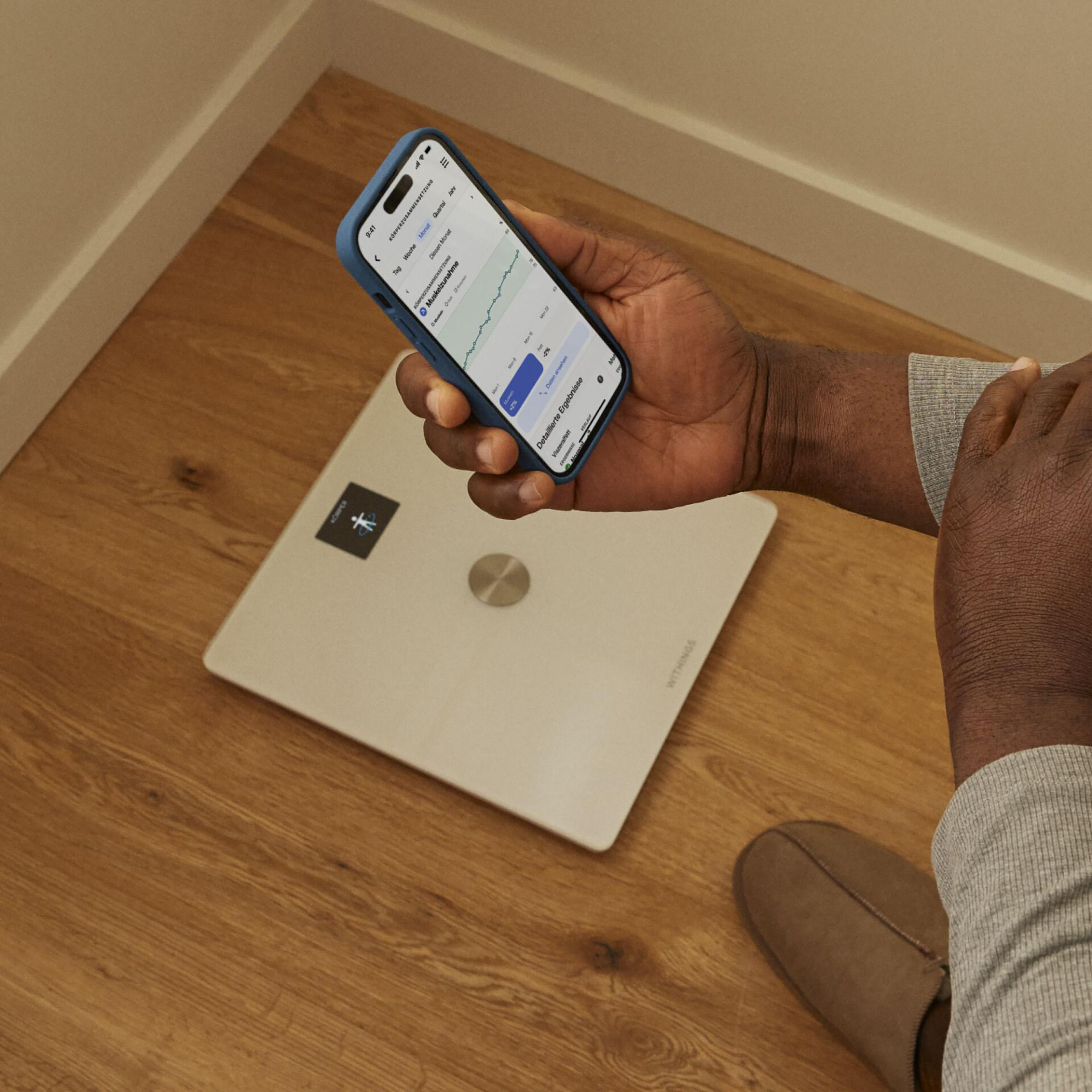 Person hält Telefon mit Gewichtsdaten neben Waage auf Holzboden.