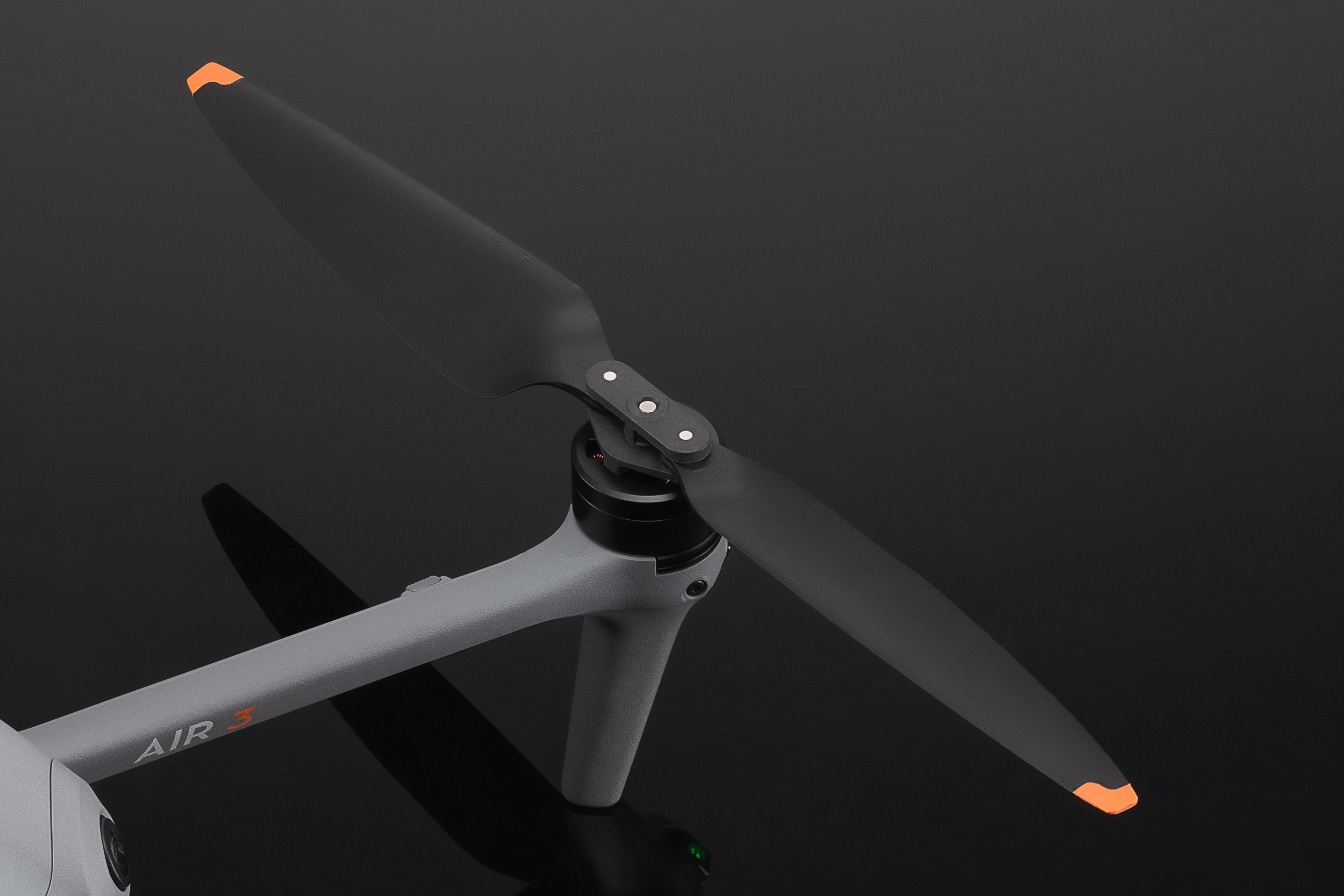Drohnenpropeller mit zwei schwarzen Blättern und orangenen Spitzen auf grauem Drohnenkörper, schwarzer Hintergrund.