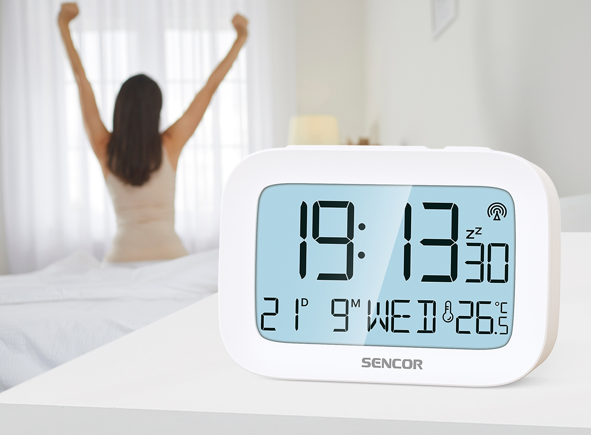 Fehér digitális ébresztőóra 19:13-mal, egy nyújtózkodó nővel a háttérben.