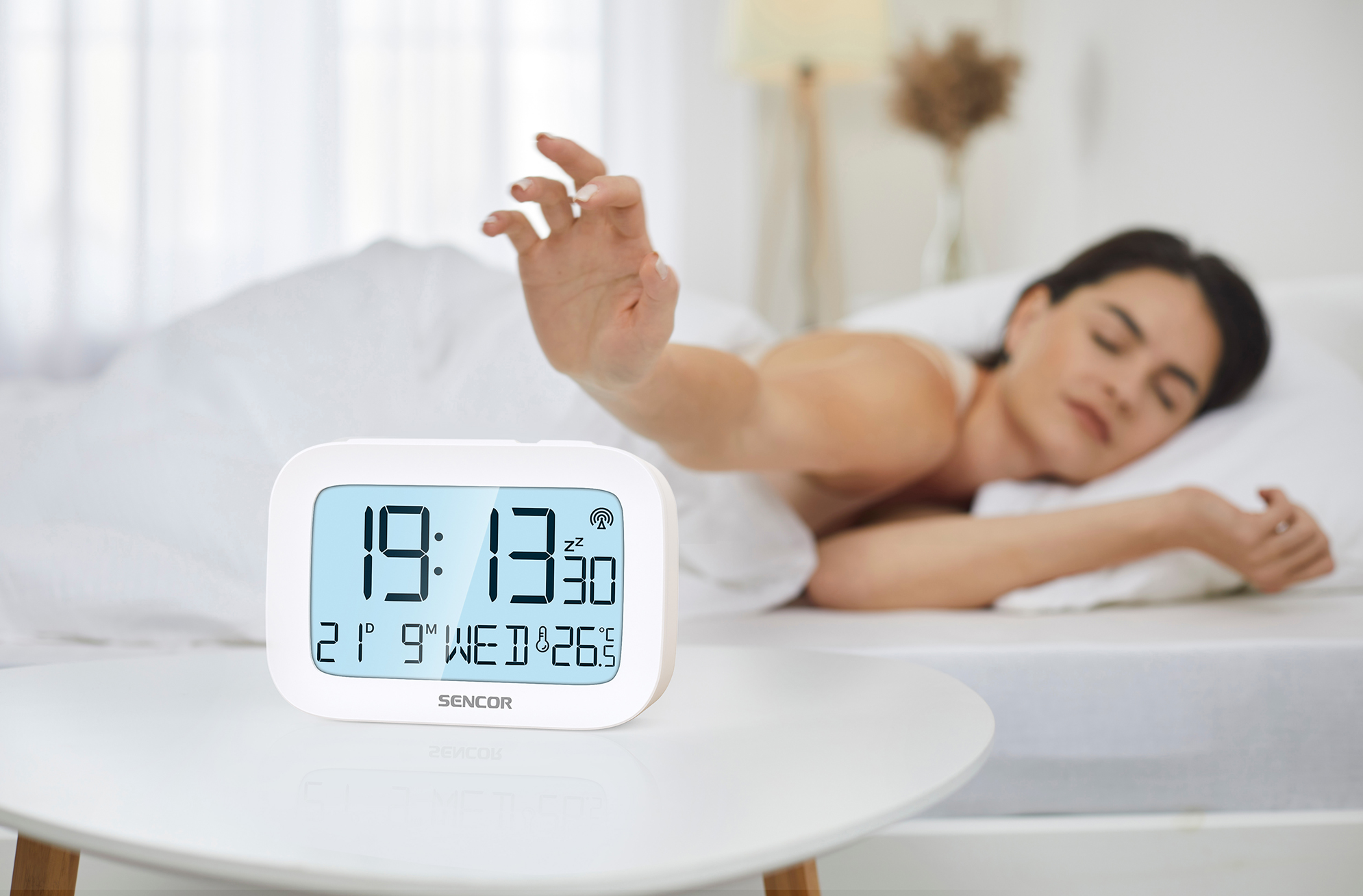 Digitális ébresztőóra az asztalon. Egy nő az ágyban nyúl felé. Ágynemű és fehér lepedők láthatók.