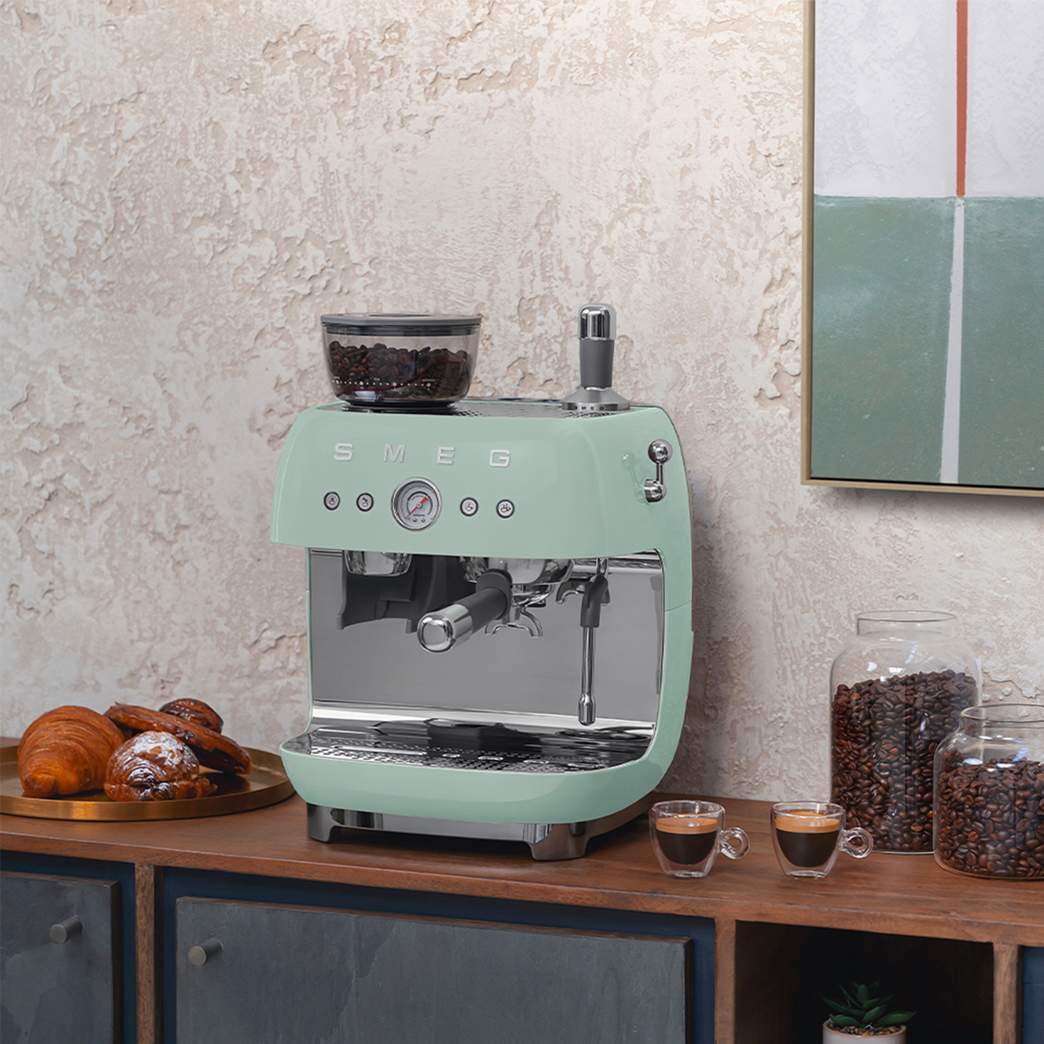 Macchina per espresso SMEG verde menta con chicchi e tazze, croissant su un bancone di legno.