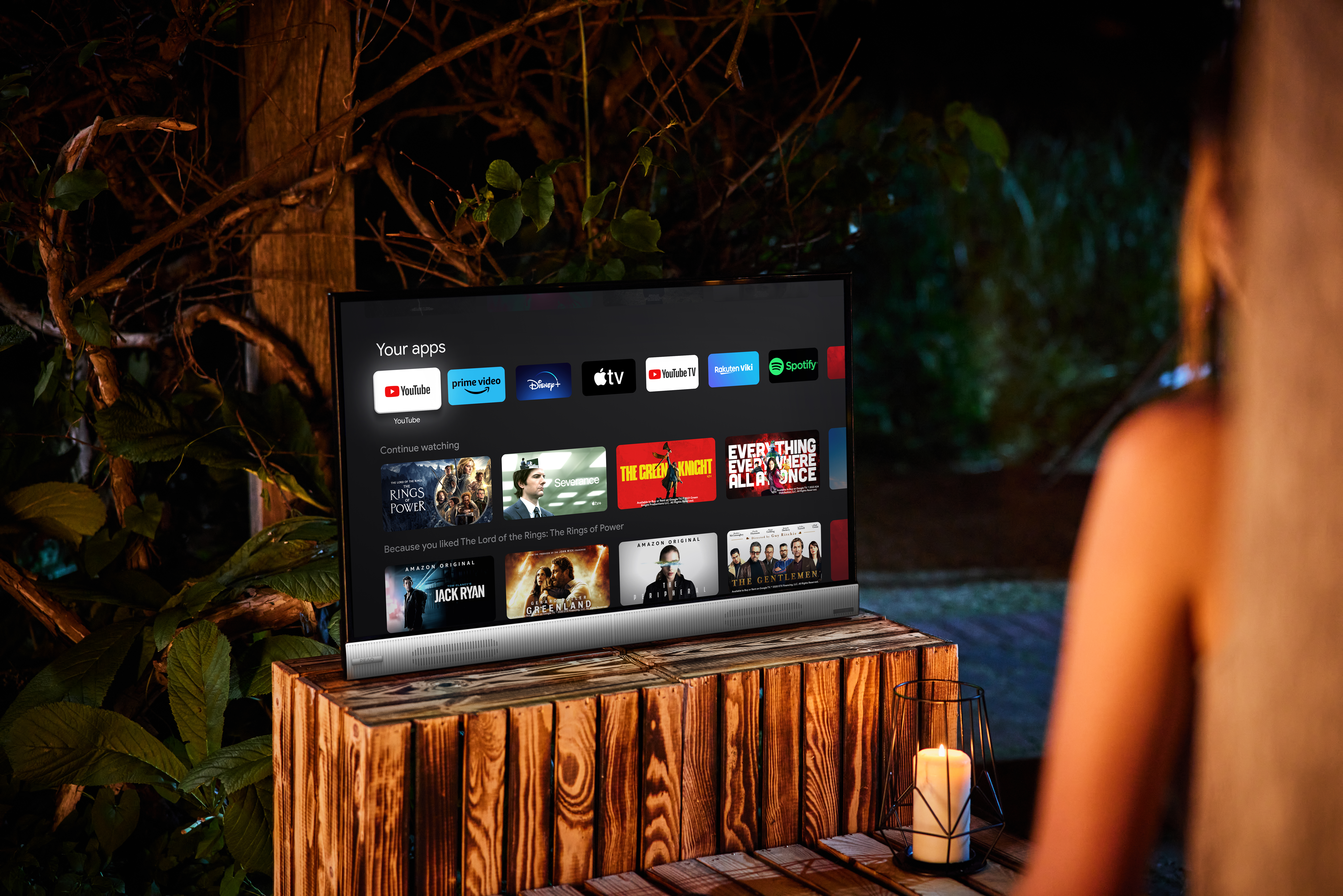 Une télévision affiche des applications et des films. Elle est posée sur une table en bois. Une personne est en arrière-plan, et une bougie est sur le côté.