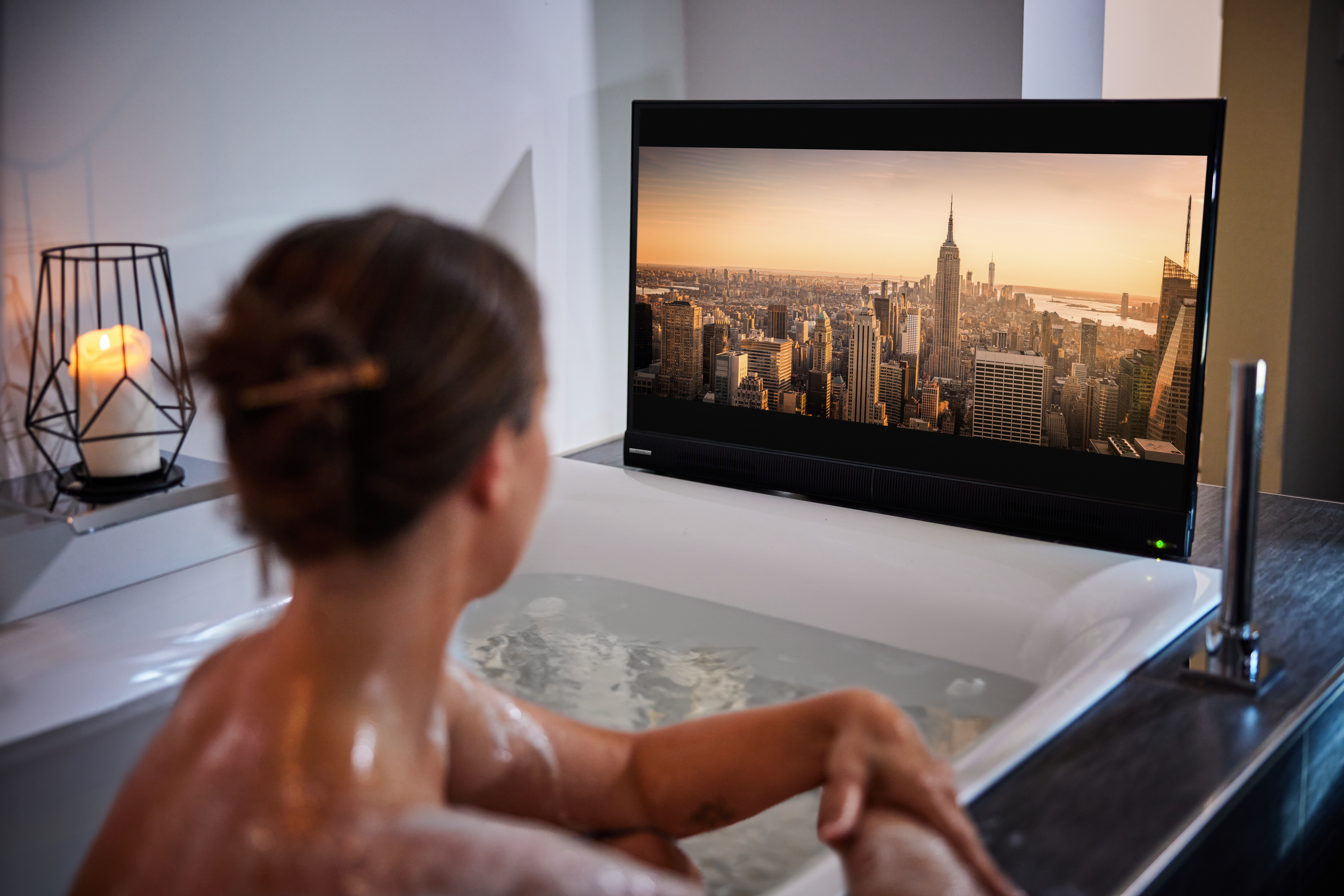 Femme dans bain regarde TV avec paysage urbain.