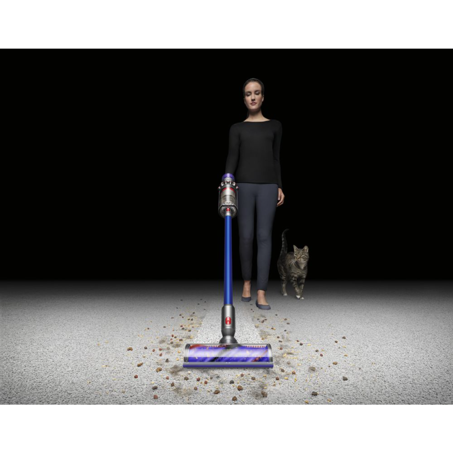 Woman vacuuming a rug with a Dyson vacuum cleaner, with a cat walking behind her.