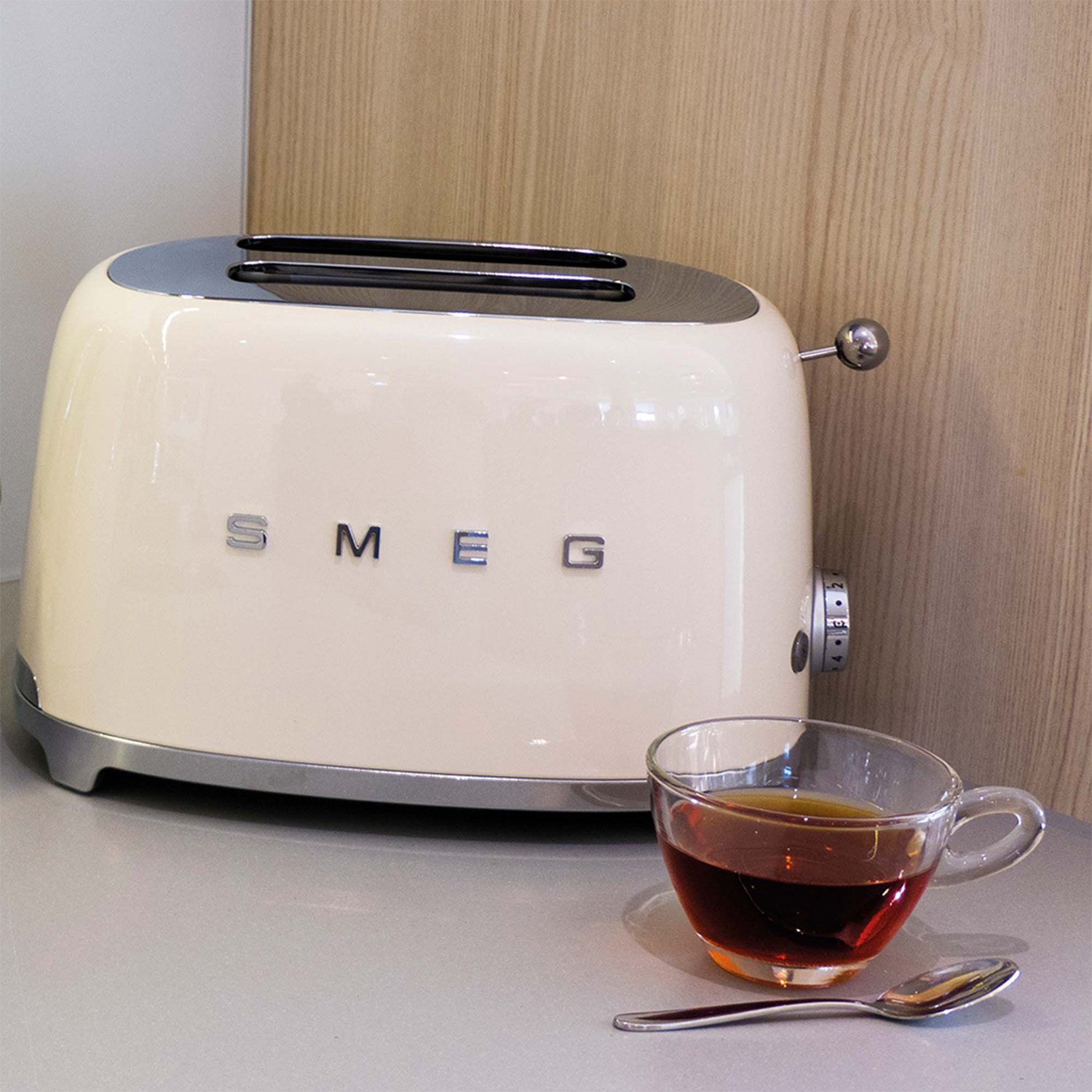 Tostapane SMEG color crema con una tazza di tè e un cucchiaio su una superficie grigia, accanto a un muro di legno.