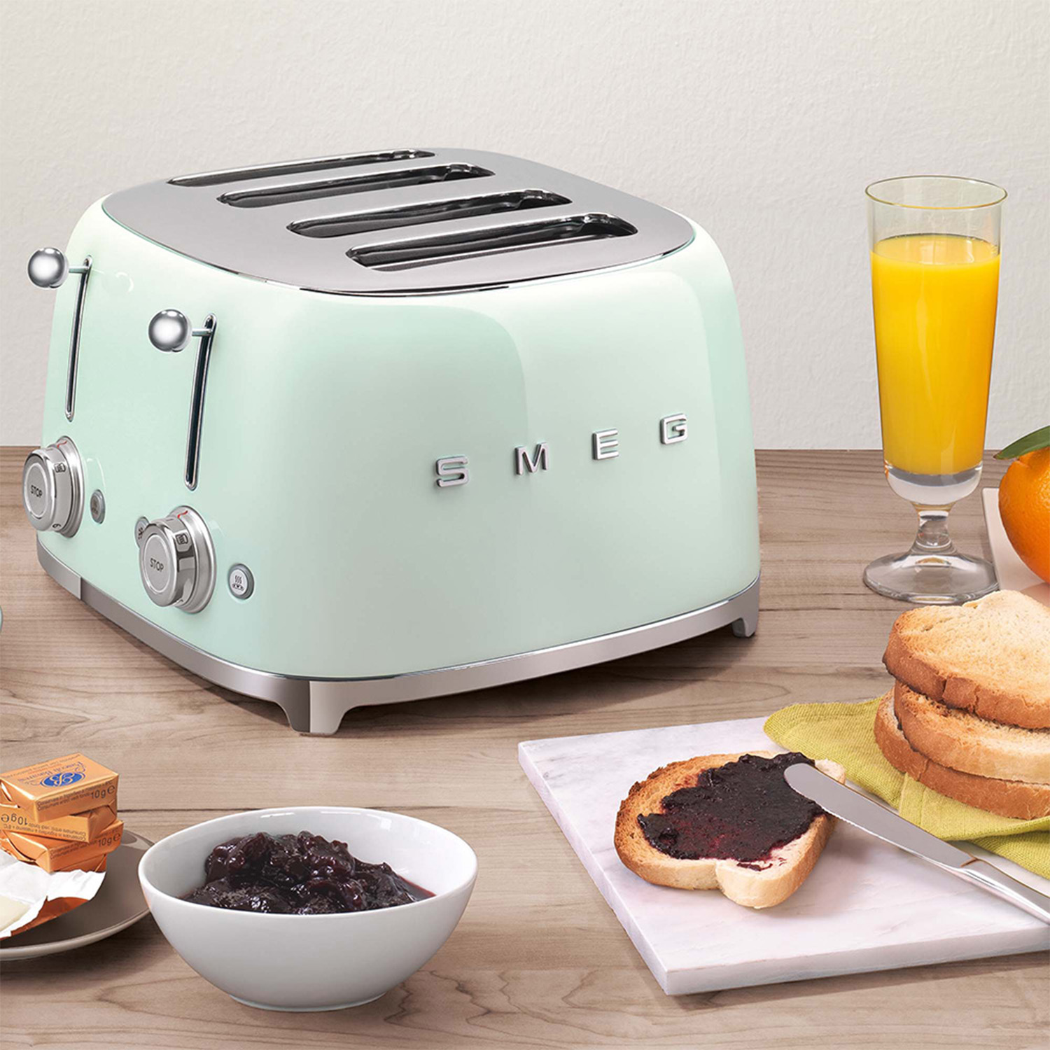 Un tostapane Smeg verde menta con articoli per la colazione su un tavolo di legno. Anche un bicchiere di succo.