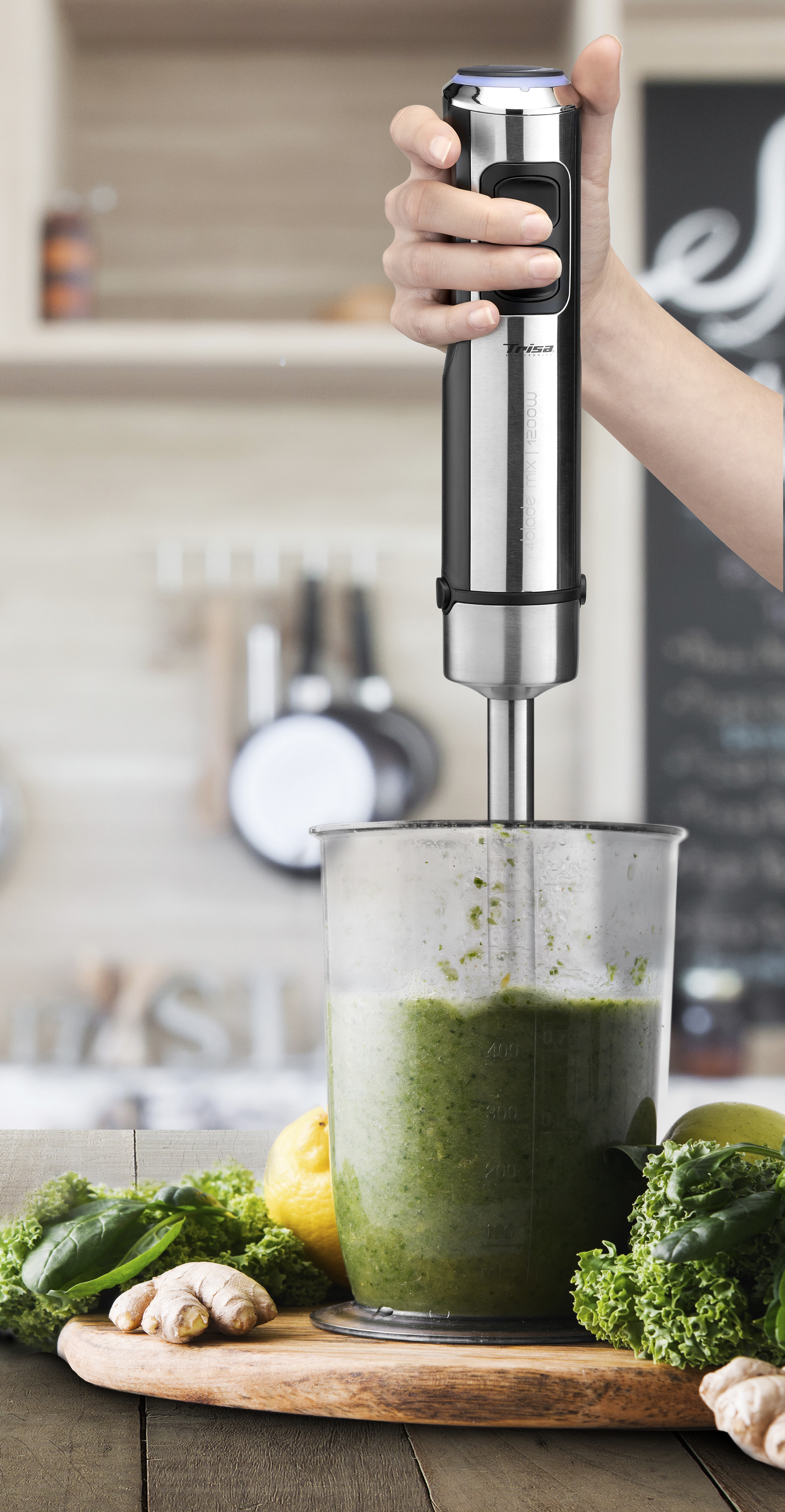 Mano che tiene un frullatore a immersione su un bicchiere di liquido verde. Tavola di legno con ingredienti.