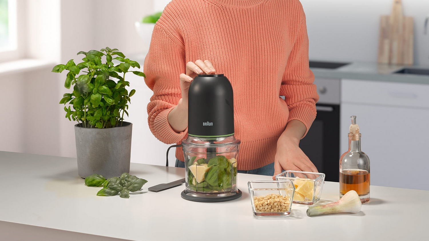 Persona haciendo pesto con un procesador de alimentos Braun, ingredientes en la mesa.