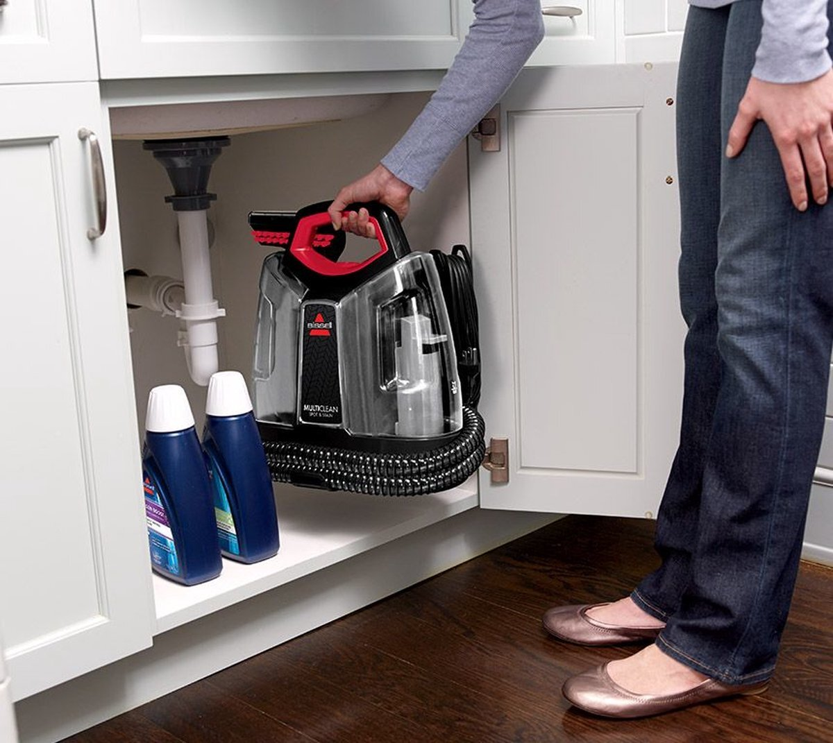 Osoba przechowuje czarno-czerwoną maszynę do czyszczenia dywanów Bissell pod zlewem.