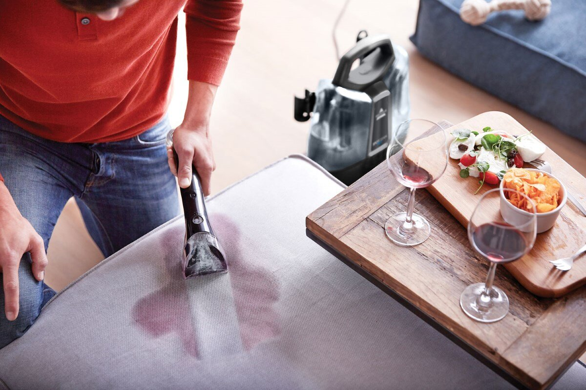 Mężczyzna czyszczący szarą sofę odkurzaczem ręcznym, z jedzeniem i winem w pobliżu.