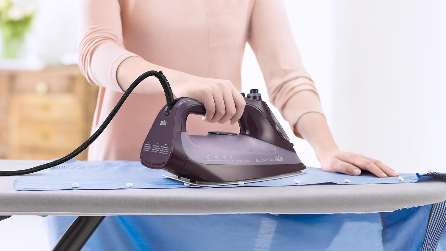 Persona planchando una camisa azul en una tabla de planchar con una plancha morada y un cable negro.