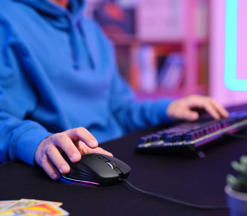 Személy fekete gamer egeret és billentyűzetet használ az asztalon, kék kapucnis pulcsiban.