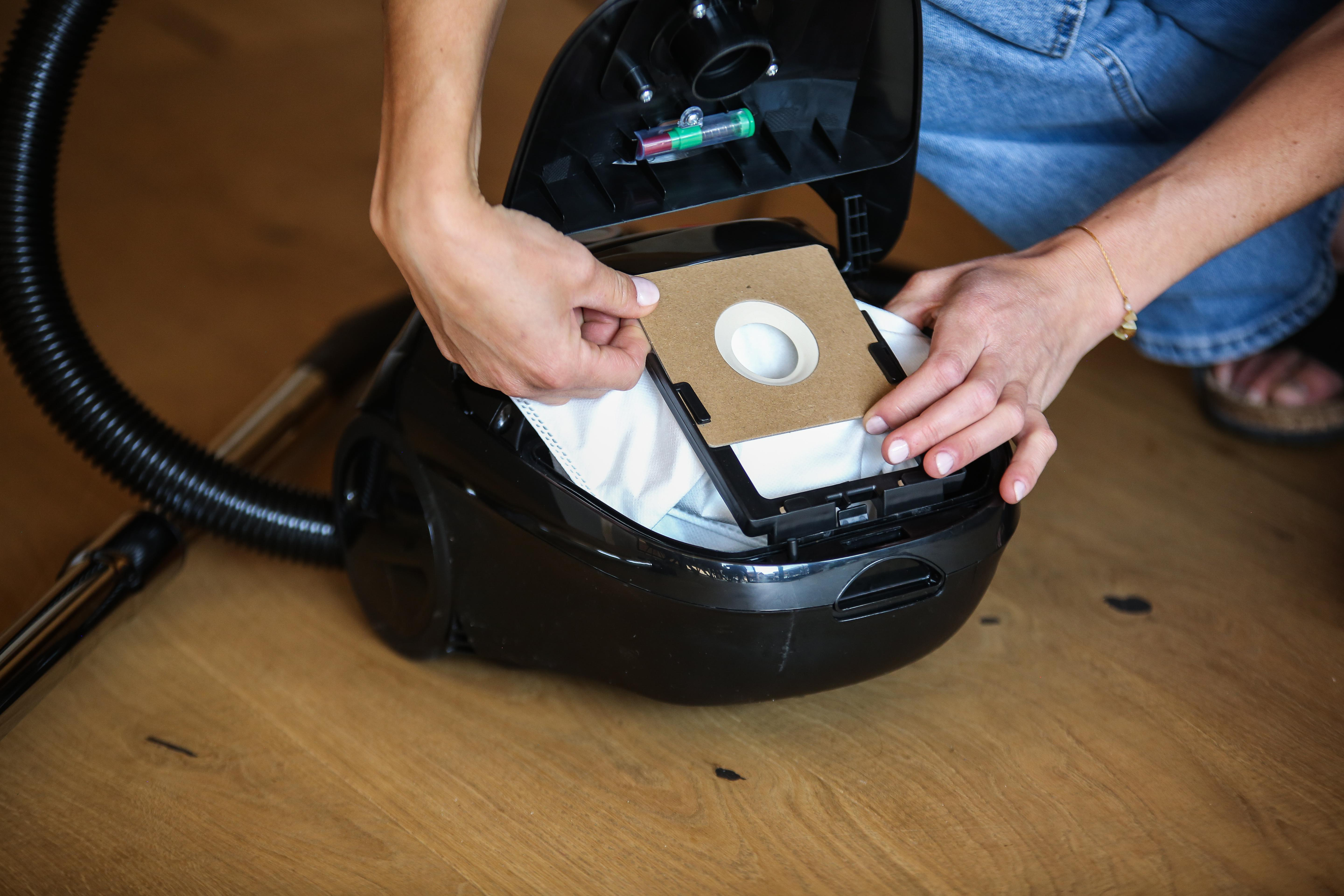 Person setzt einen neuen Staubsaugerbeutel ein. Schwarzer Staubsauger auf Holzboden.