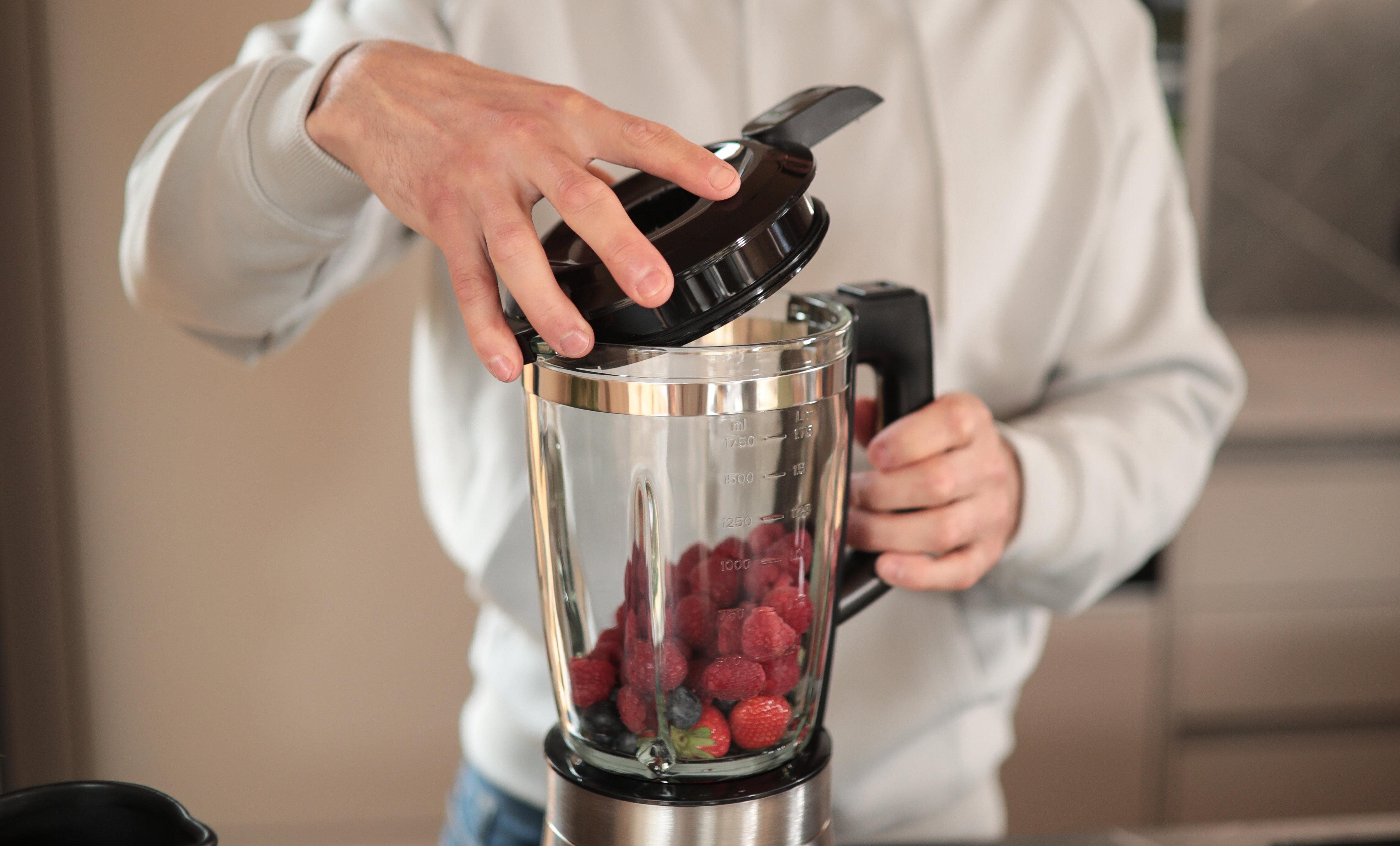 Person öffnet einen Mixer. Beeren befinden sich im Glasbehälter.