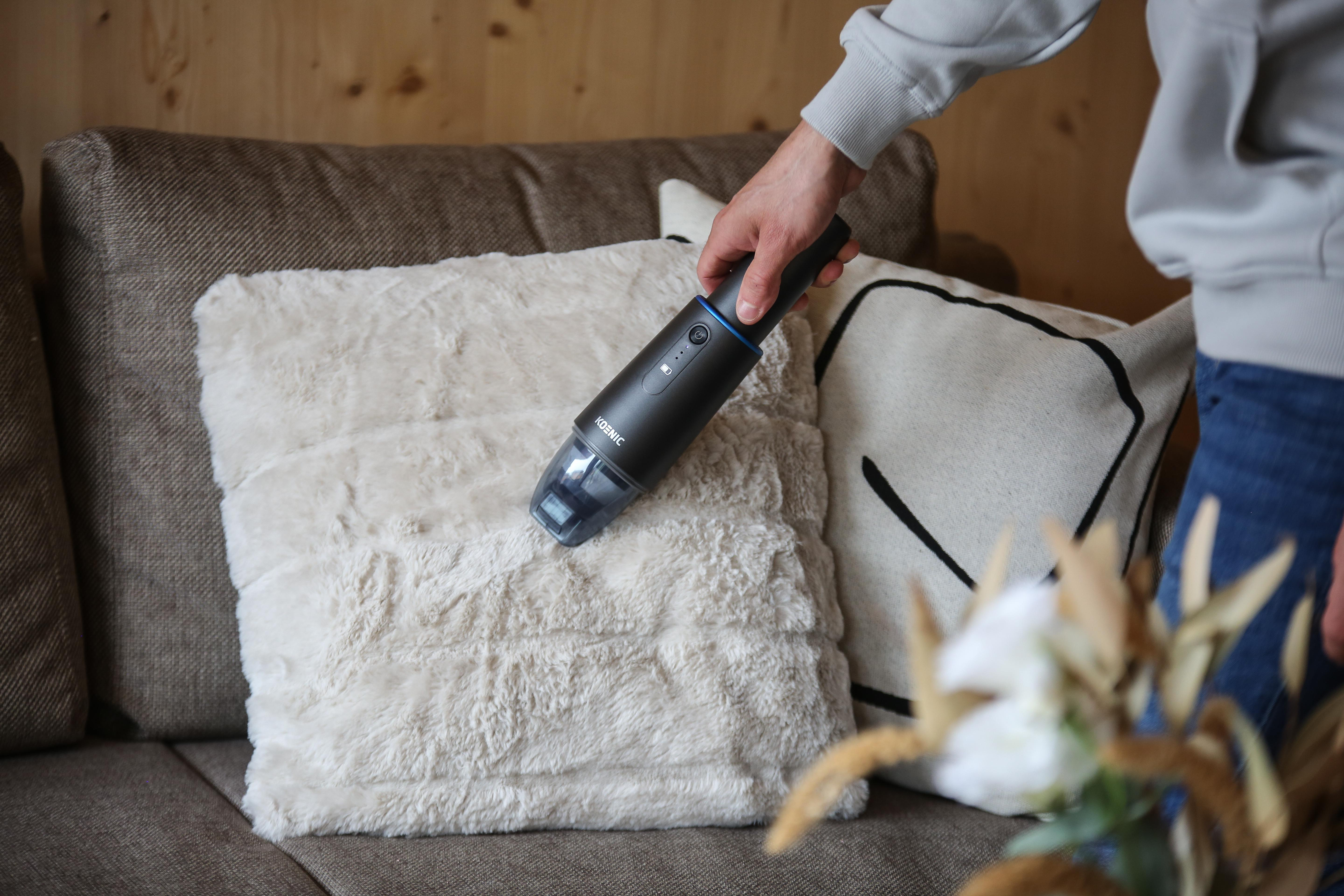 Eine Person, die einen flauschigen cremefarbenen Kissen auf einem braunen Sofa mit einem Handstaubsauger absaugt.