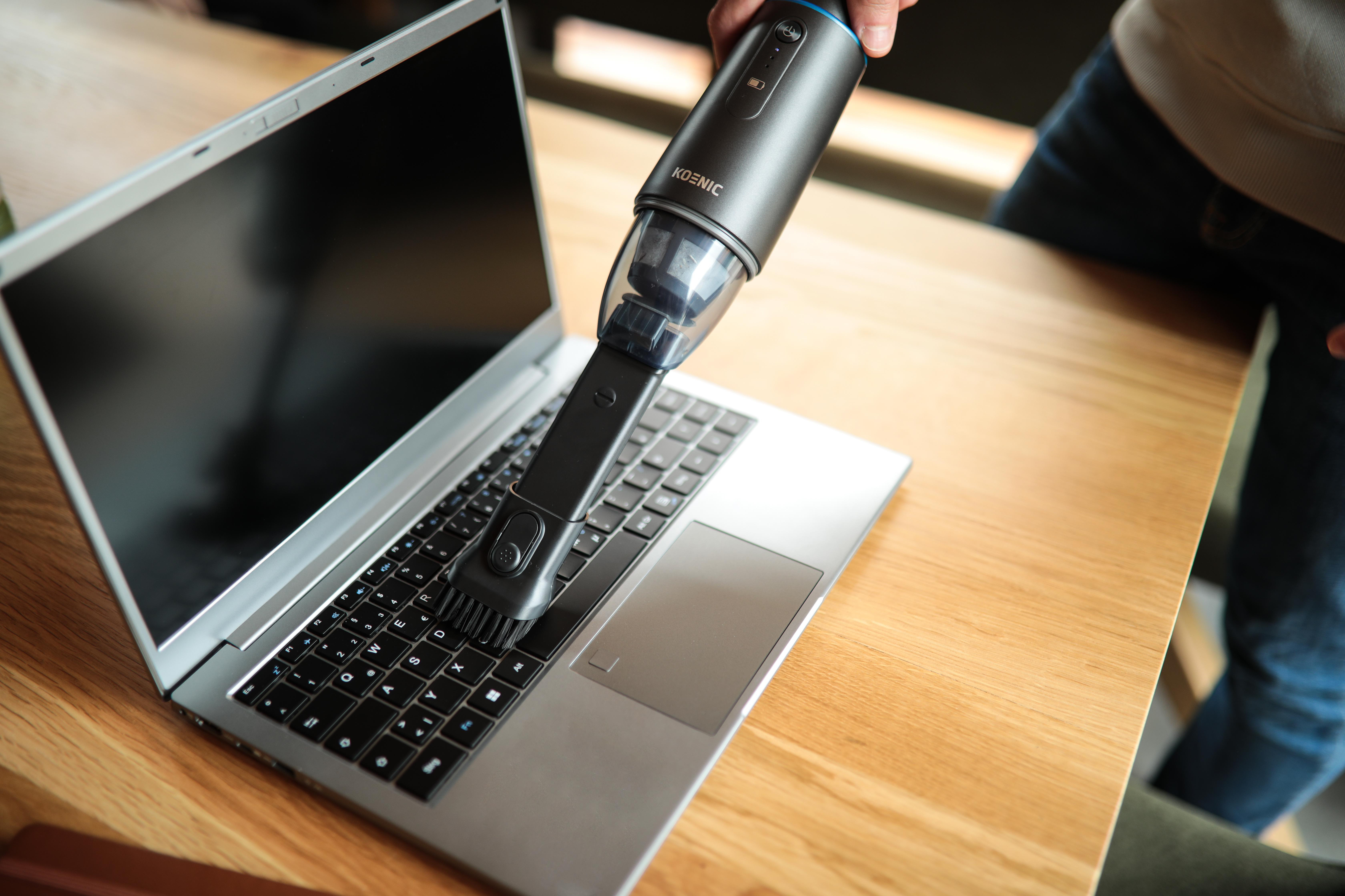 Eine Person saugt eine Laptop-Tastatur. Der Staubsauger ist schwarz und der Laptop steht auf einem Holztisch.