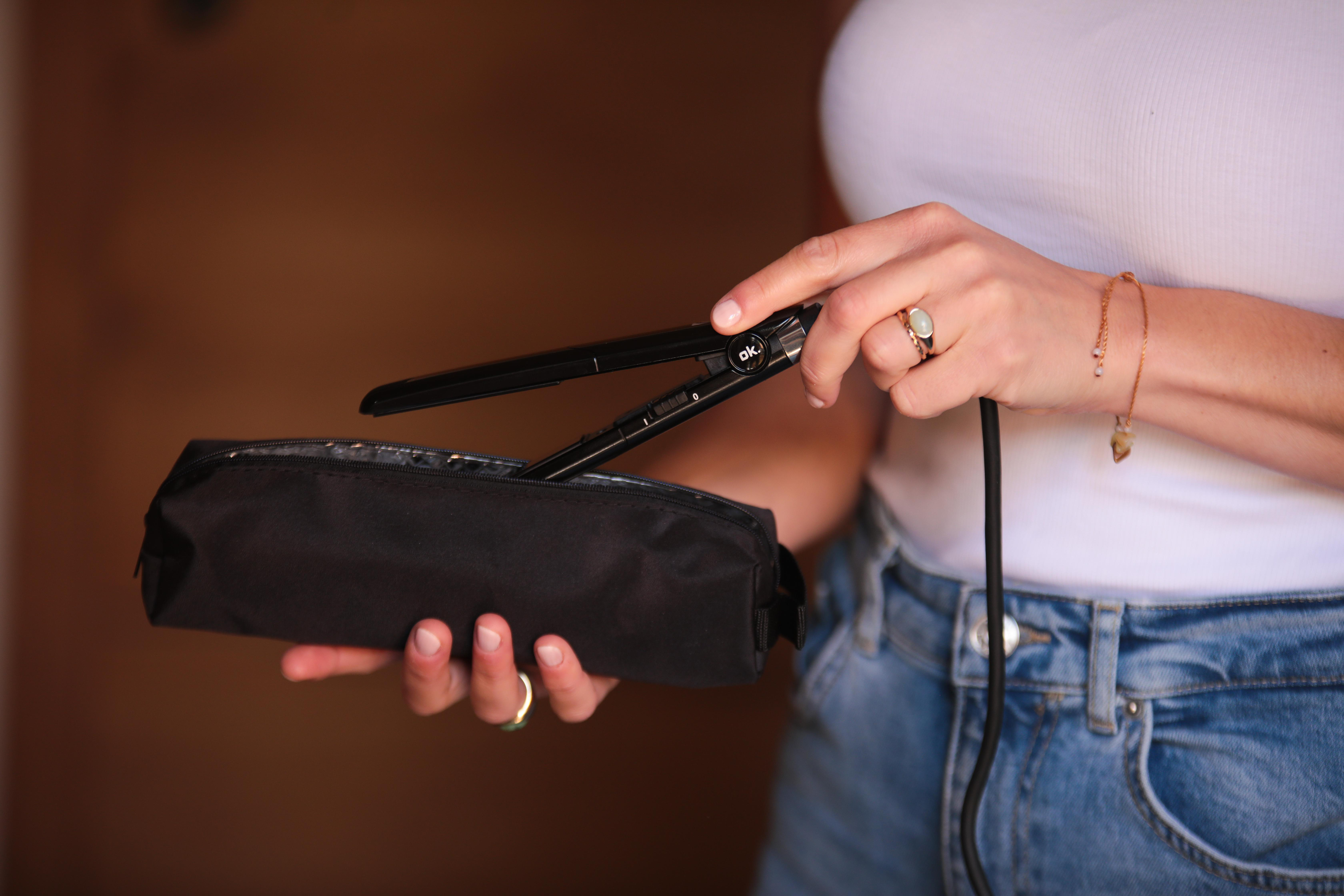 Eine Frau hält ein schwarzes Glätteisen und eine schwarze Tasche. Sie trägt Jeans und ein weißes Hemd.