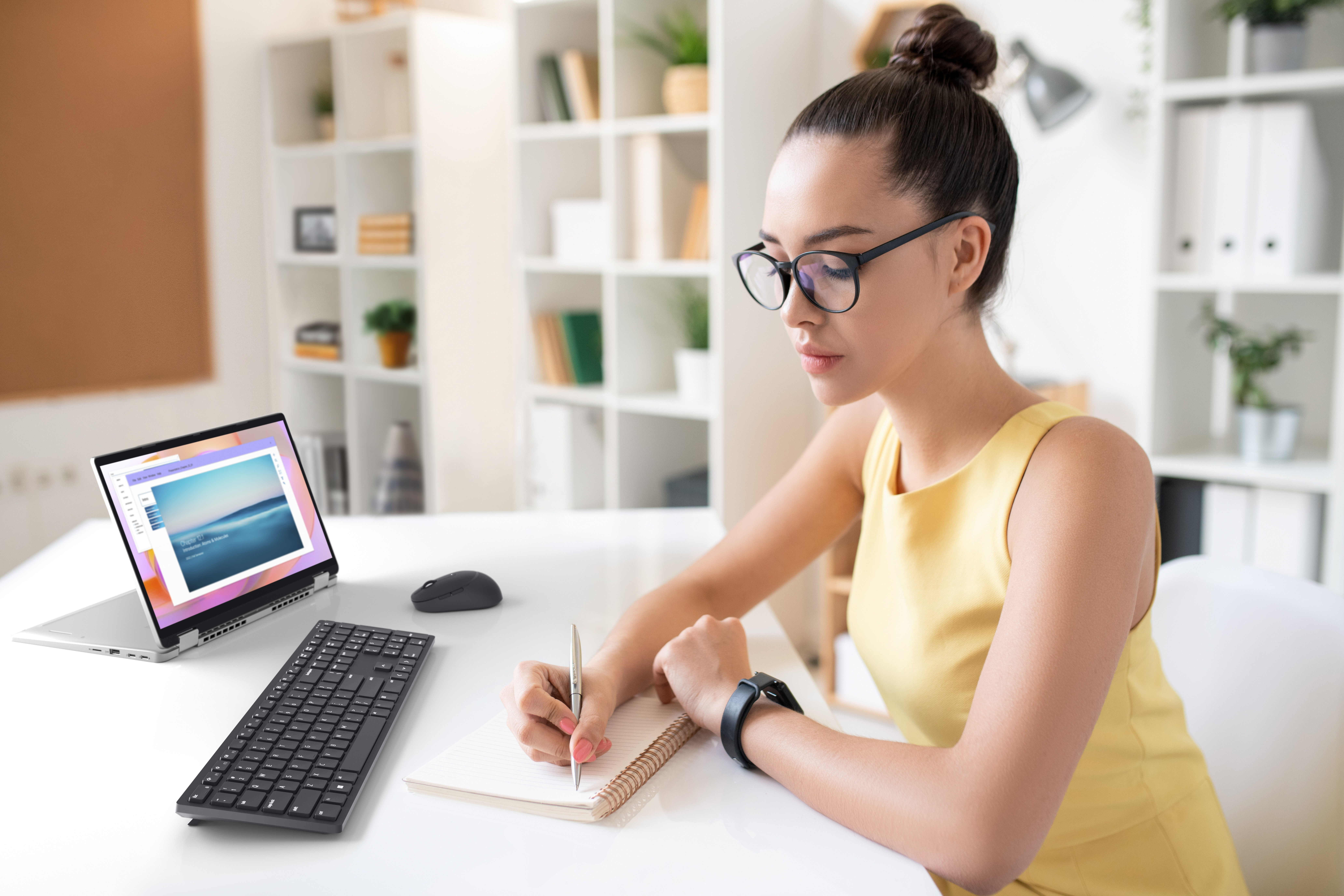 Frau mit Brille schreibt in ein Notizbuch am Schreibtisch. Laptop und Tastatur sind vor ihr.