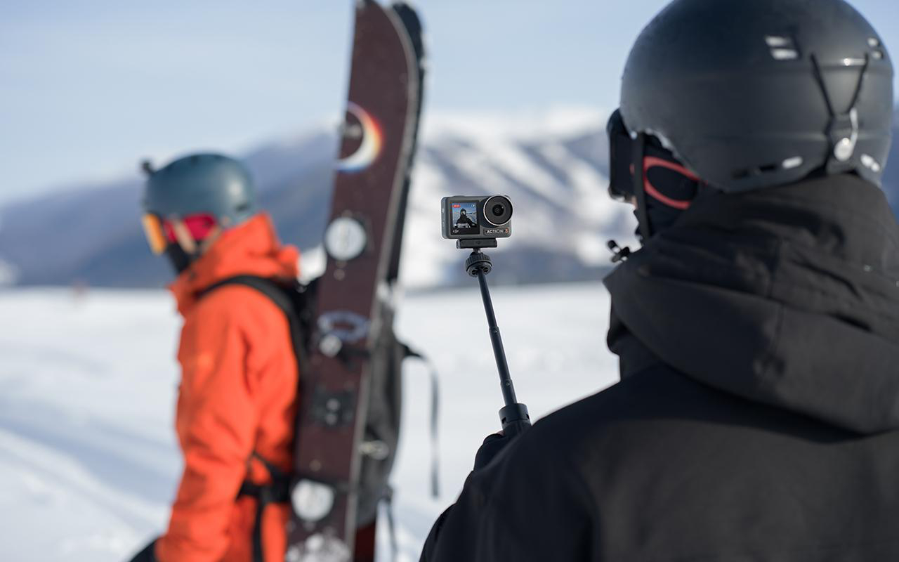 Zwei Skifahrer. Einer hält eine Kamera an einer Stange. Verschneiter Berg im Hintergrund.