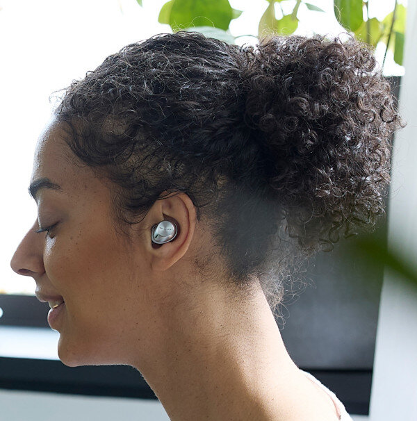 Kobieta z kręconymi włosami nosząca srebrną słuchawkę. Zielone rośliny w tle.