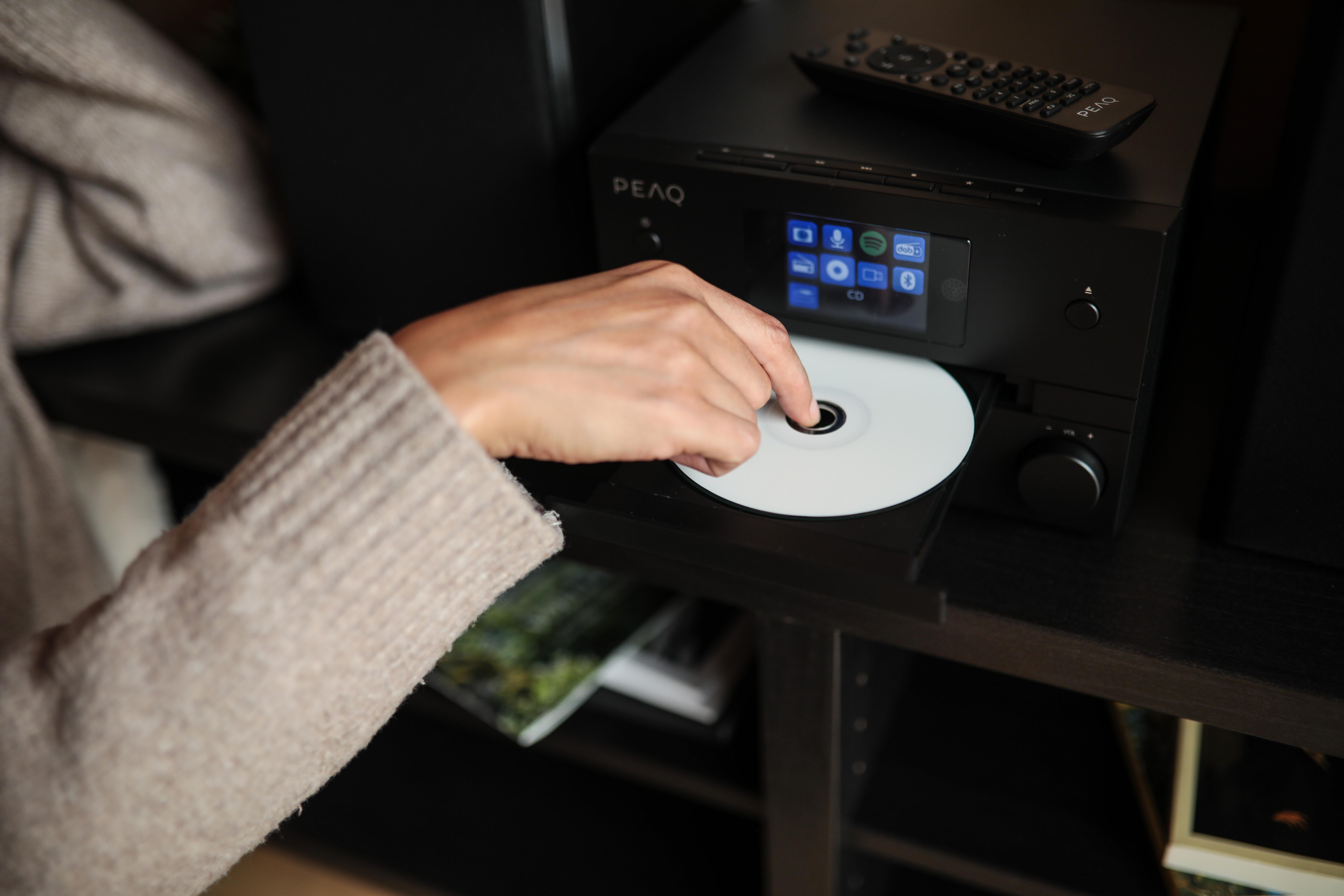 Eine Person legt eine CD in ein schwarzes Audiosystem mit Bildschirm ein.