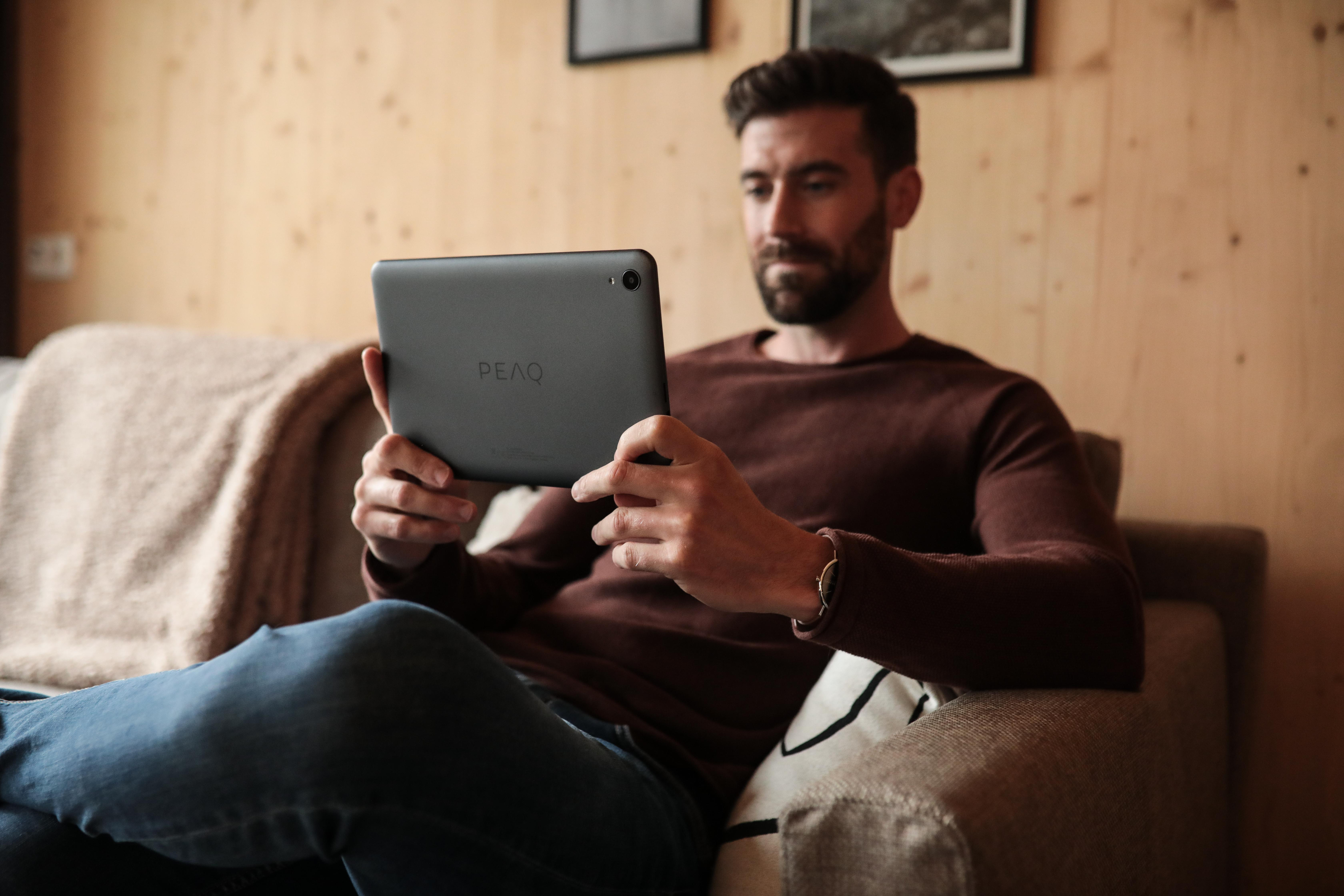 Ein Mann sitzt auf einem Sofa und hält ein Tablet. Er trägt einen braunen Pullover und Jeans und schaut auf den Bildschirm.