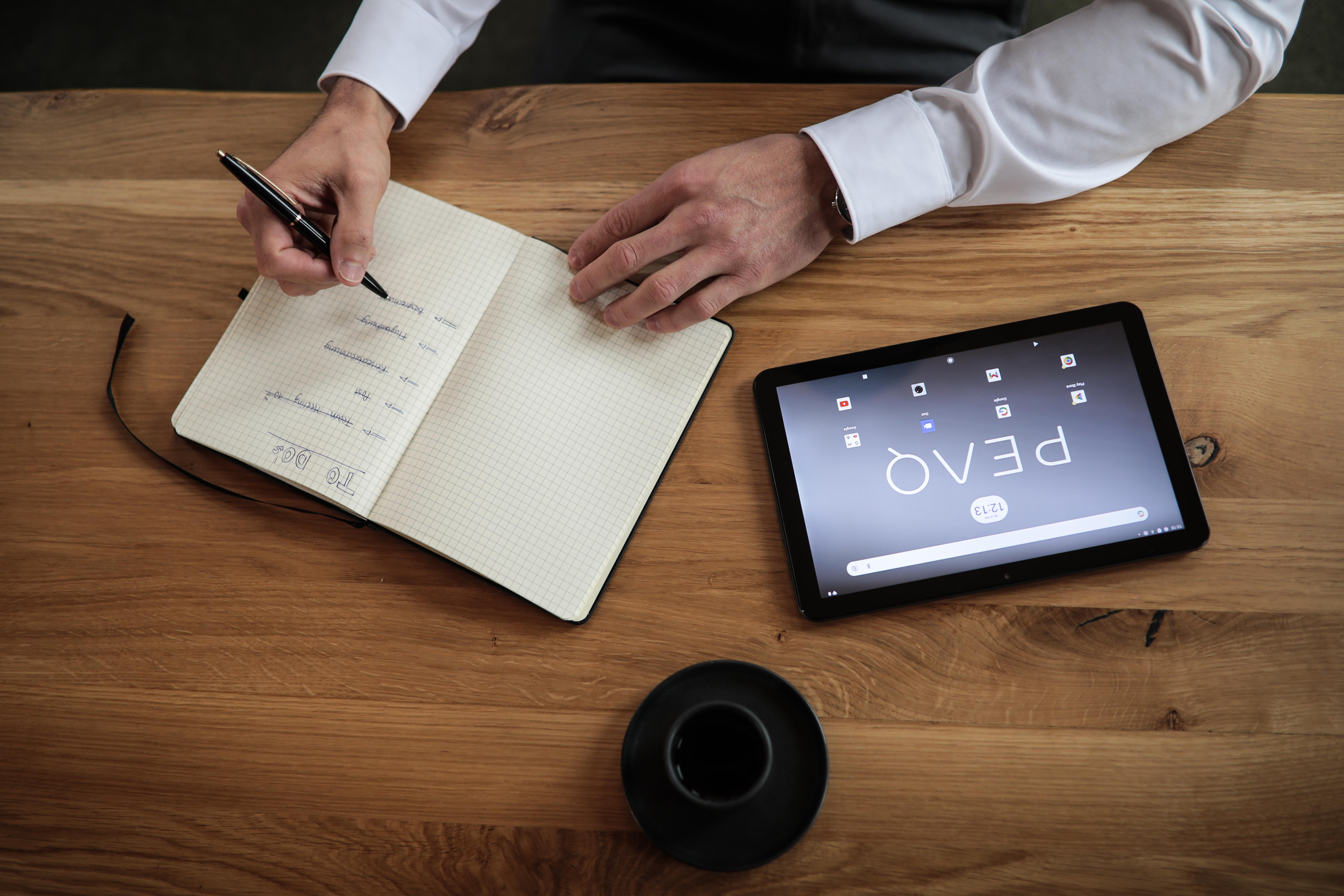 Person schreibt in ein Notizbuch, ein Tablet und eine Kaffeetasse auf einem Holztisch. Tablet zeigt Apps.