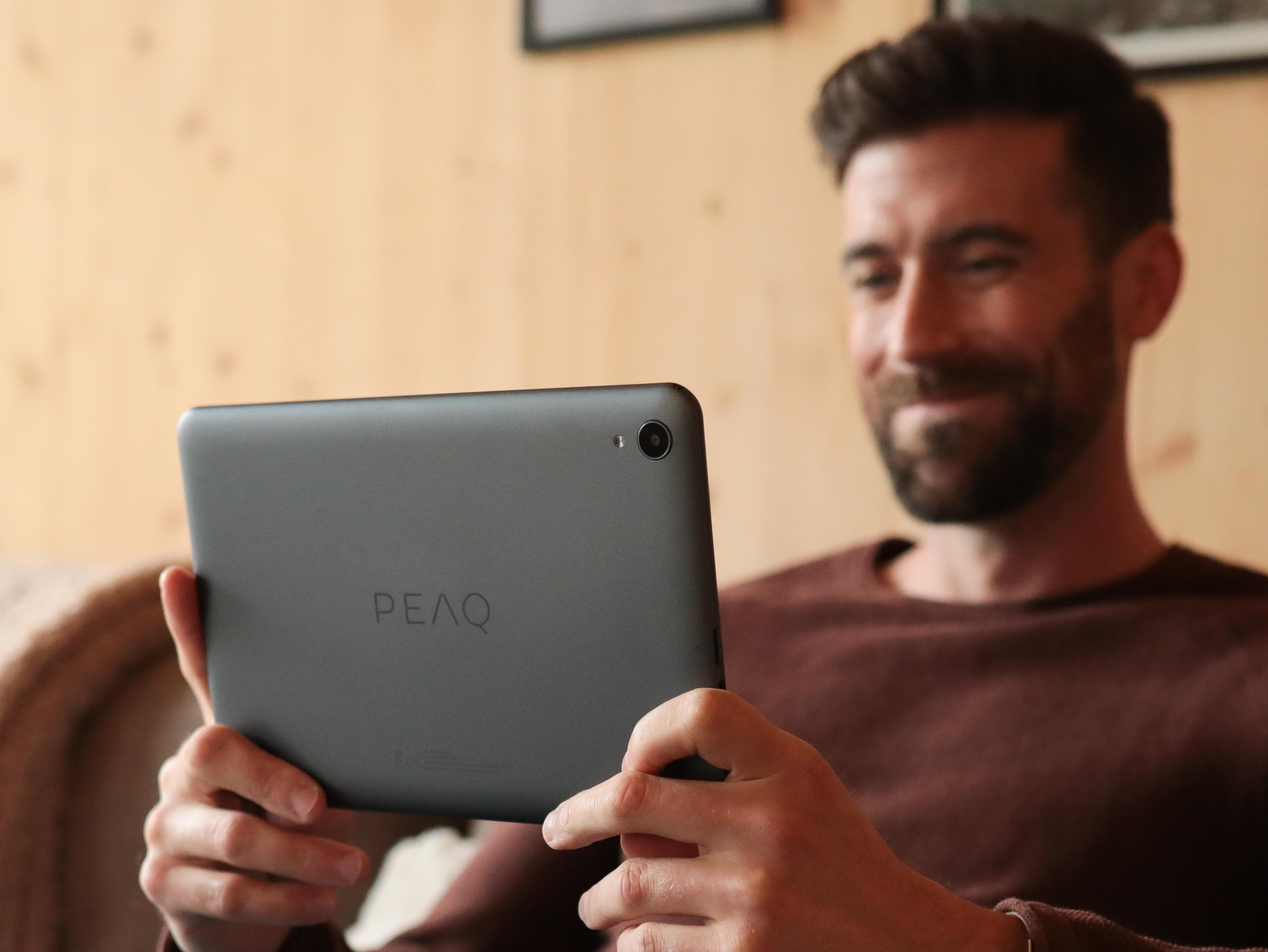 Mann lächelt, hält ein graues Tablet. Das Tablet zeigt das Wort PEAQ.