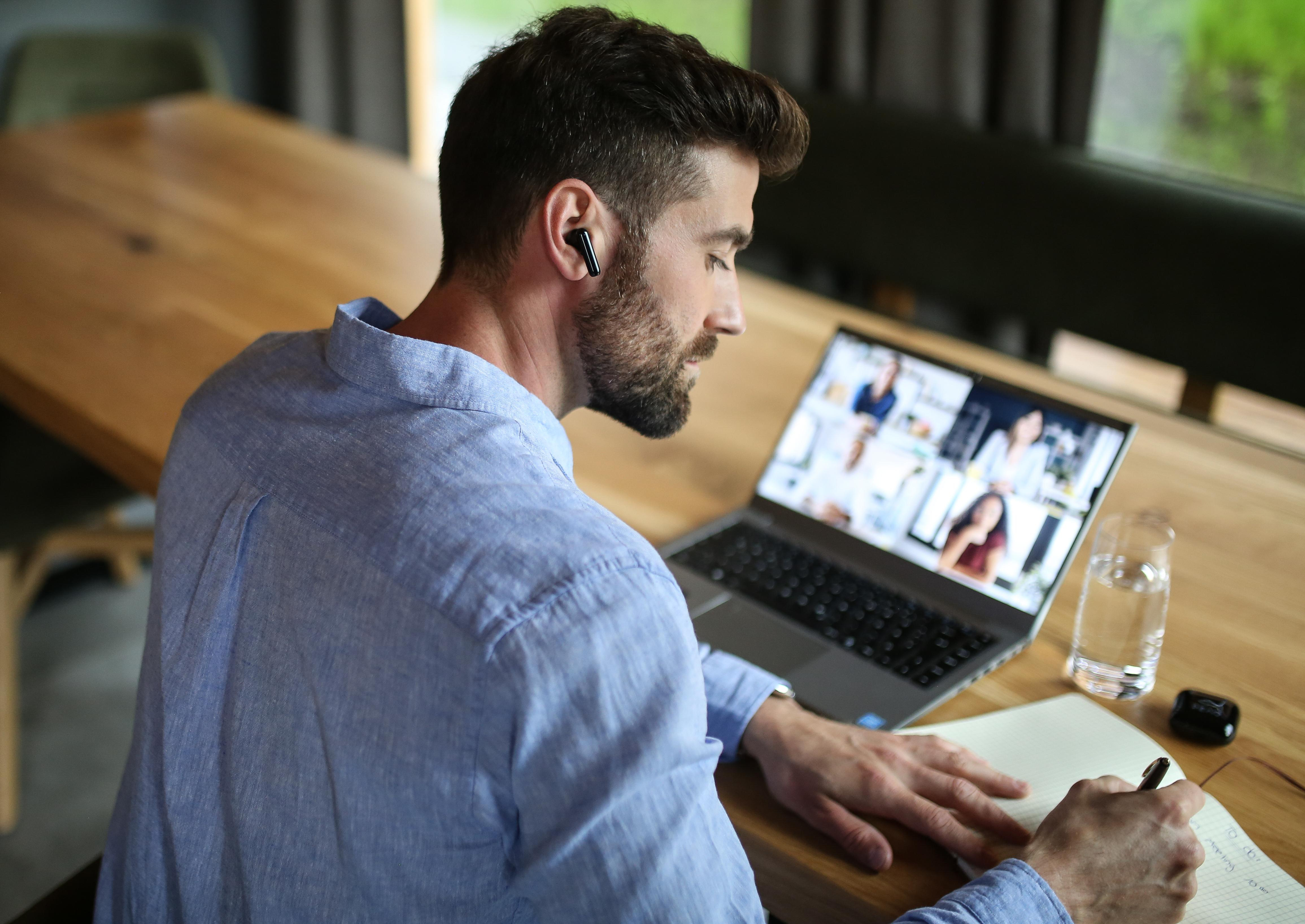 Ein Mann in blauem Hemd telefoniert per Videoanruf auf seinem Laptop, während er in ein Notizbuch schreibt.