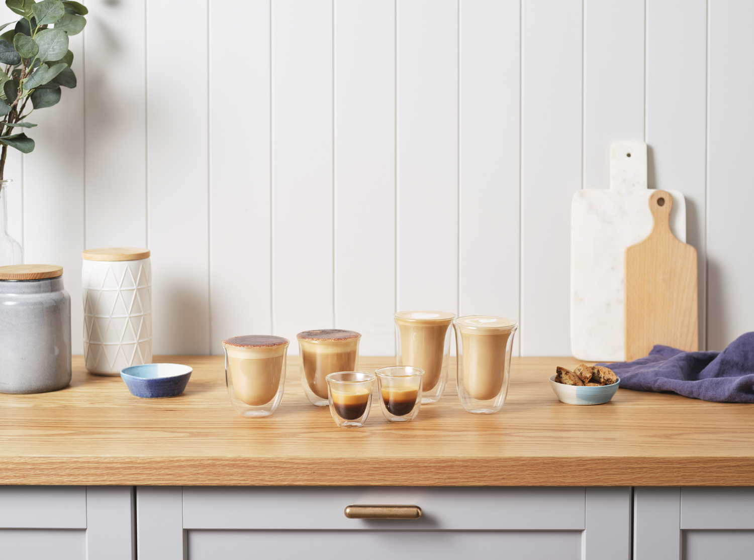 Bebidas de café en vasos de vidrio sobre una encimera de madera, con una tabla de cortar y otros utensilios.
