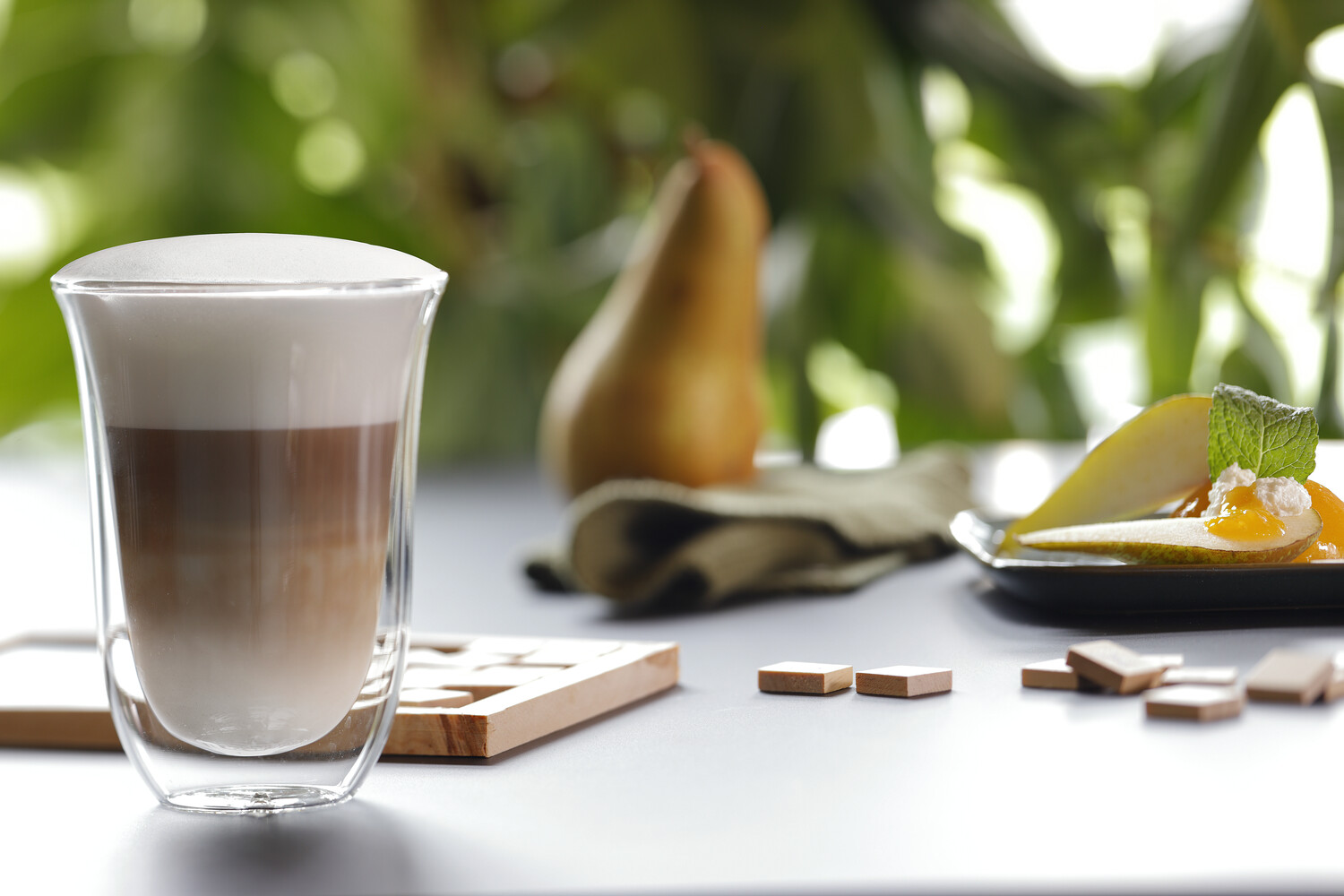 Un latte macchiato en una taza de vidrio con una pera y trozos de chocolate en la mesa.