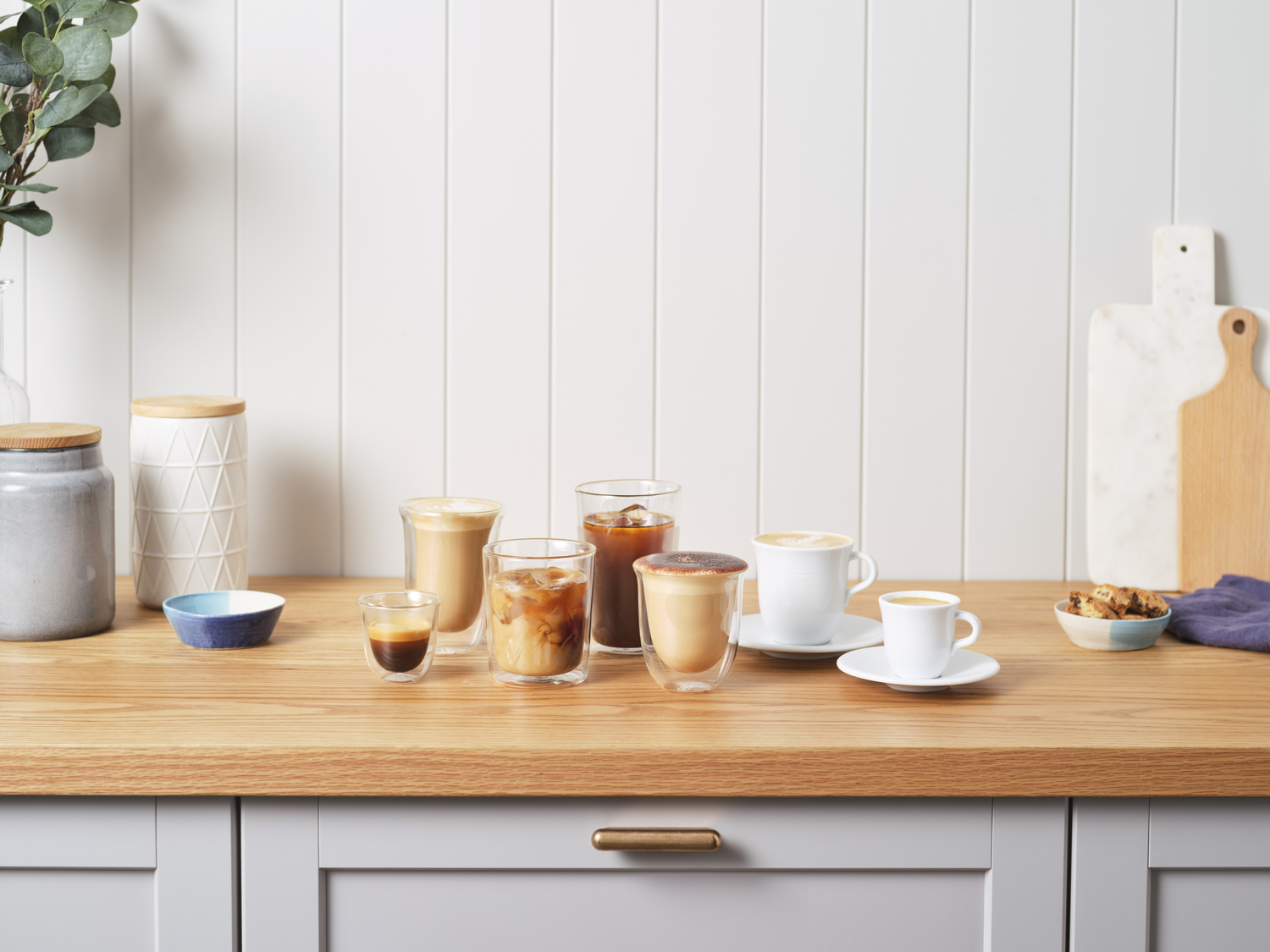 Bebidas de café en varios vasos sobre una encimera de madera. El fondo es una pared blanca.