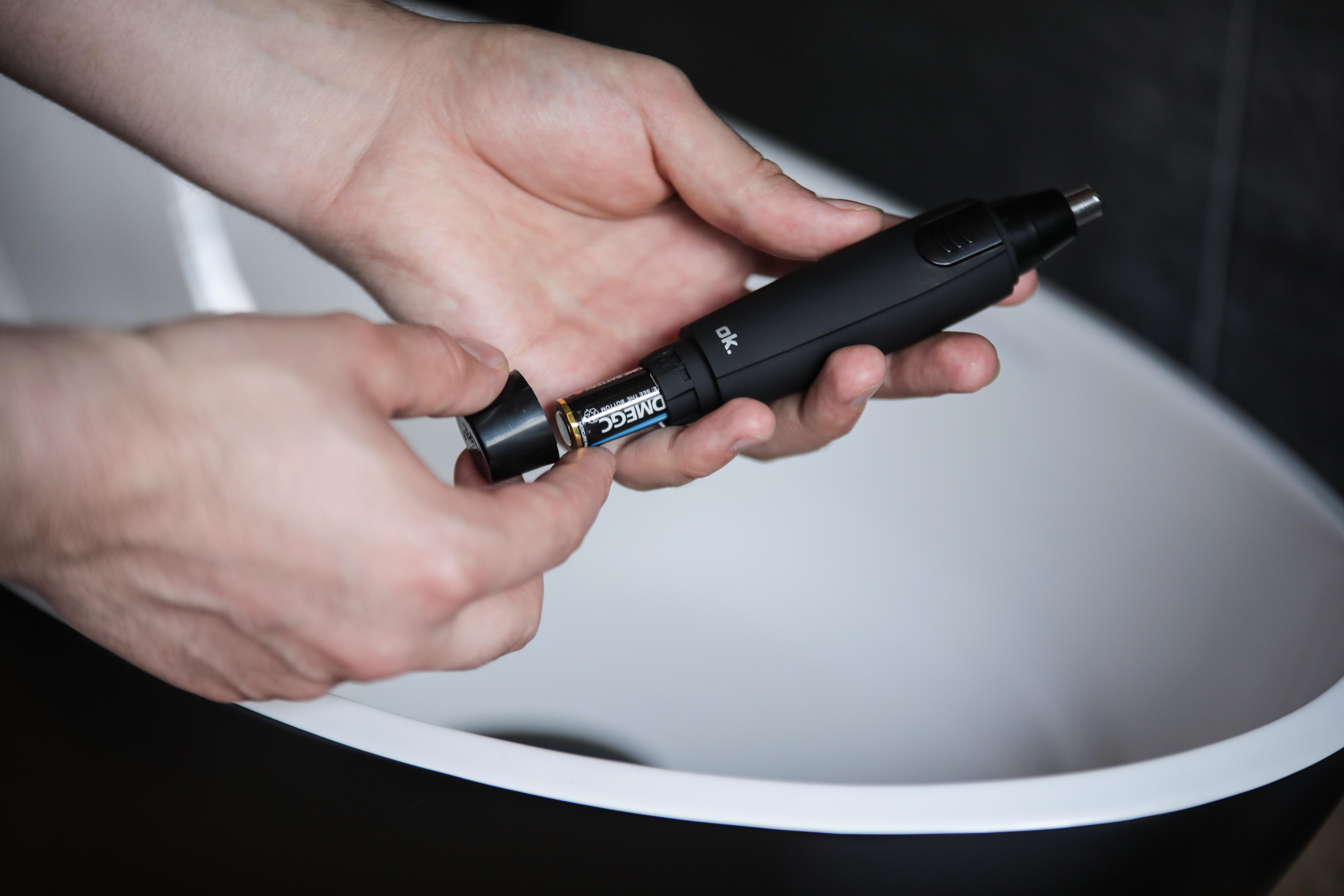 Hände, die eine Batterie in einen schwarzen Nasenhaartrimmer einsetzen, in der Nähe eines weißen Waschbeckens.