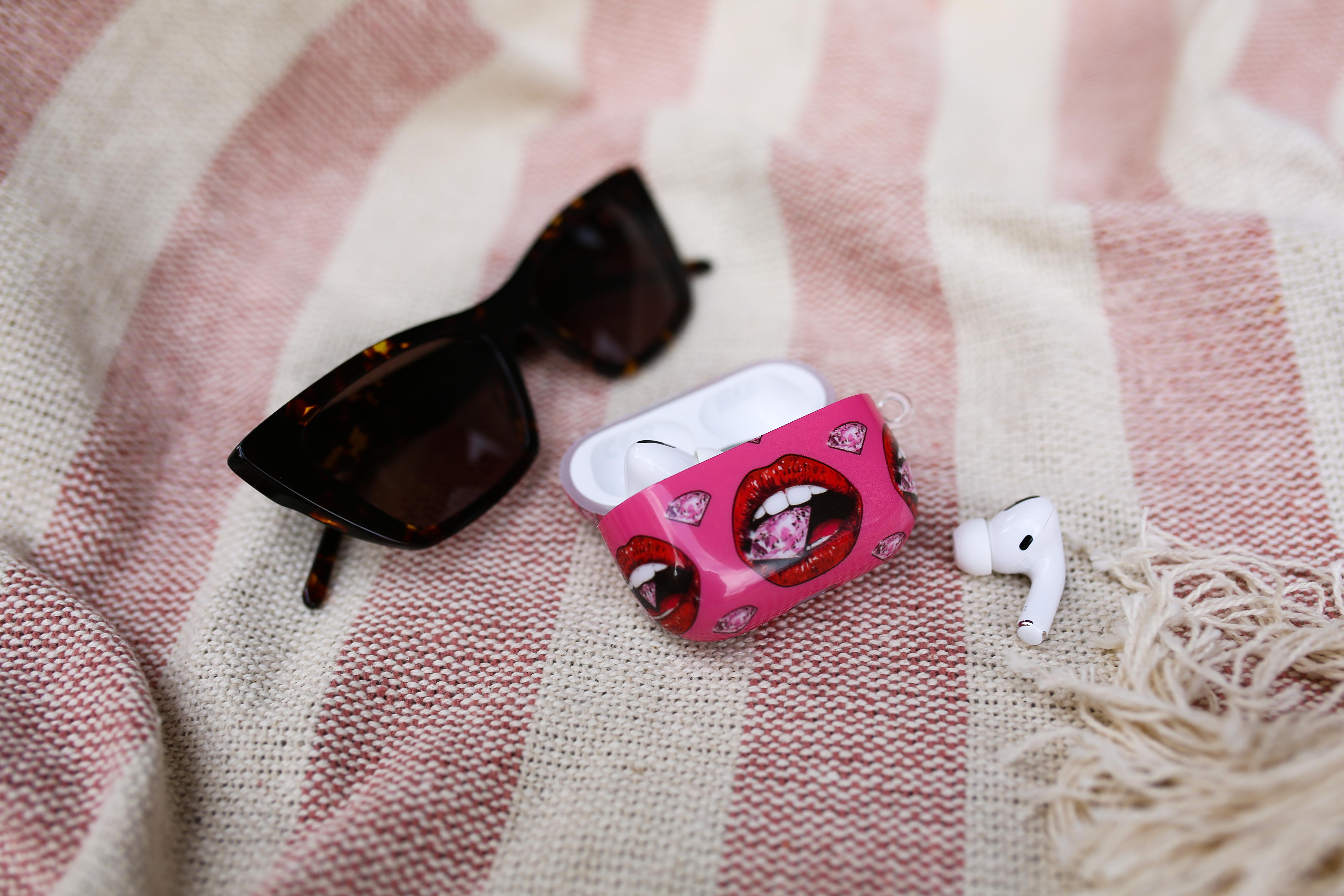 Kopfhörer-Etui mit rosa Lippen-Design. Sonnenbrille auf einer gestreiften Decke. Weißer Ohrhörer.