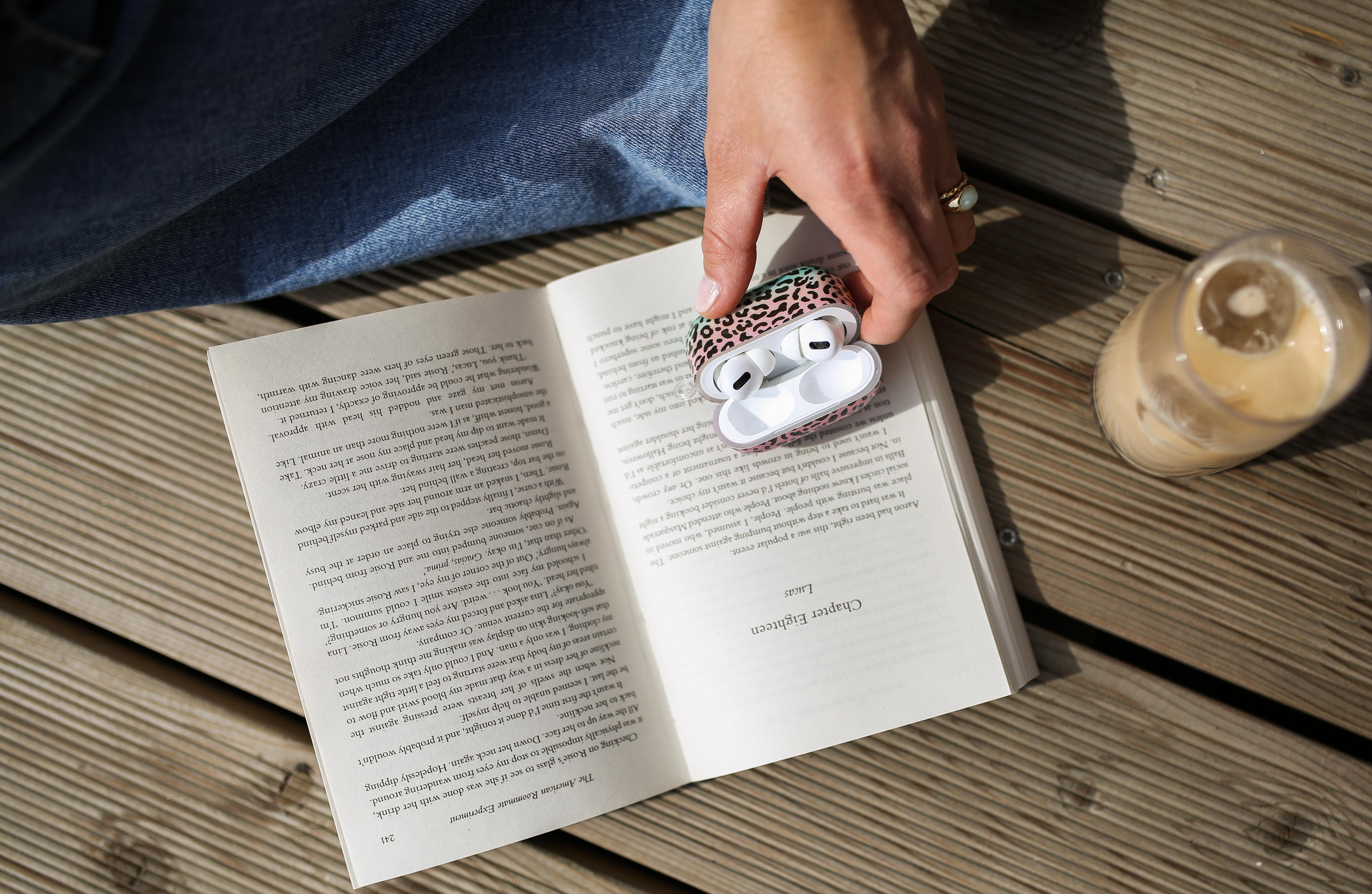 Person hält AirPods-Etui und ein Buch. Eiskaffee daneben. Holzdeck.