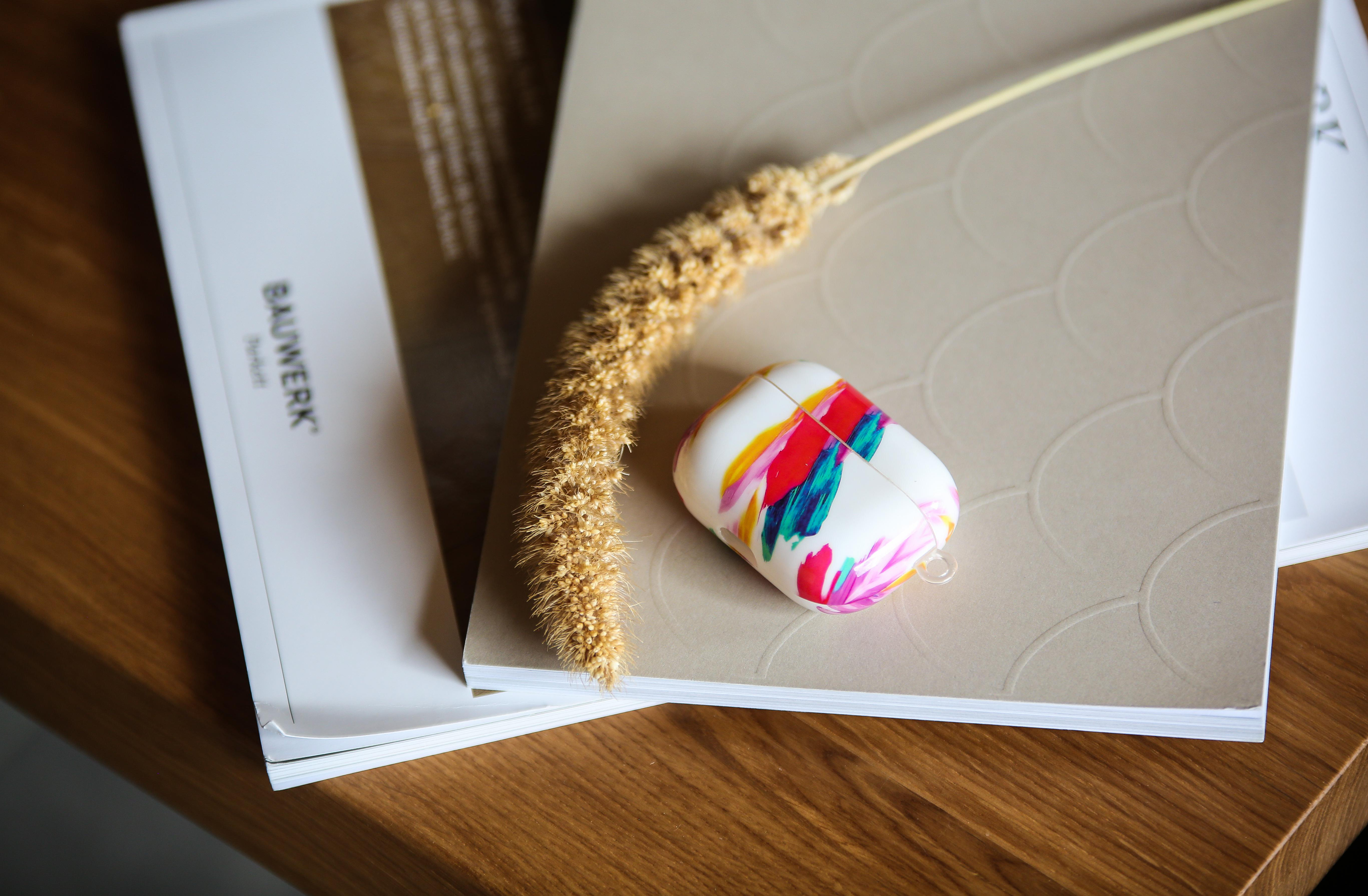 AirPods-Hülle mit bunten Farbspritzern, auf einem Notizbuch mit Stift liegend.