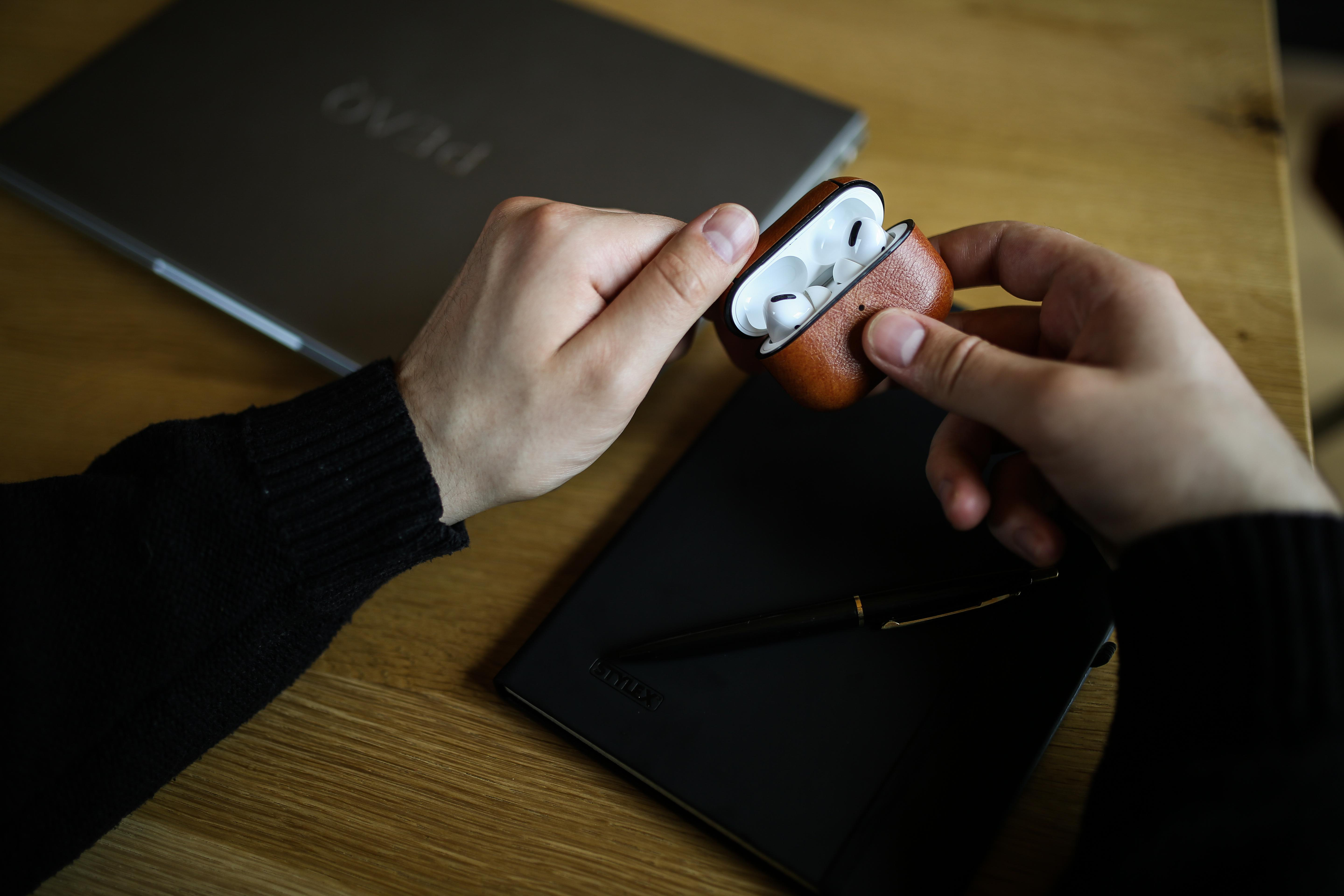 Eine Person, die ein braunes Leder-AirPods-Etui hält und einen Laptop und ein Notizbuch betrachtet.
