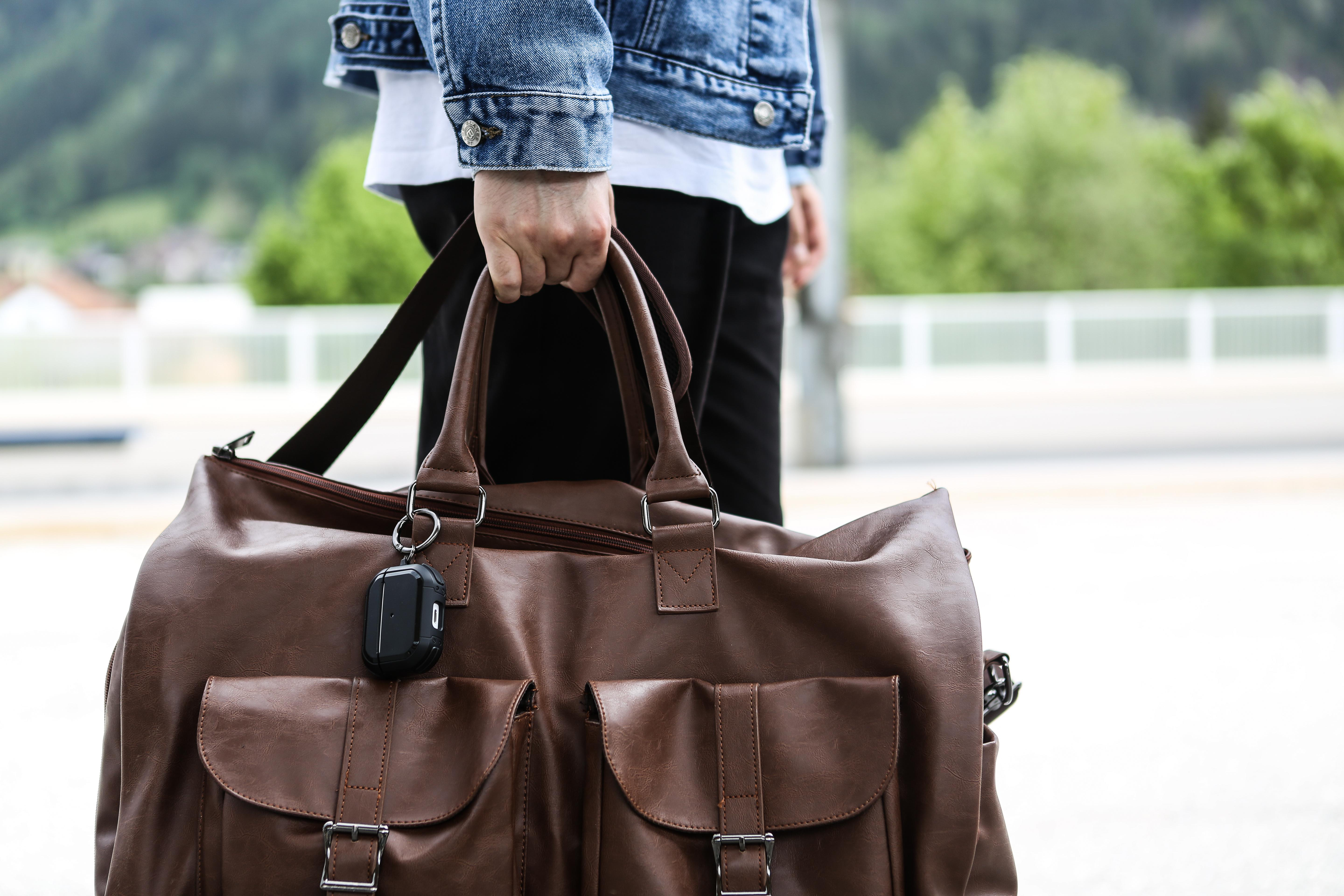 Person hält eine braune Reisetasche mit einem schwarzen Etui am Reißverschluss.