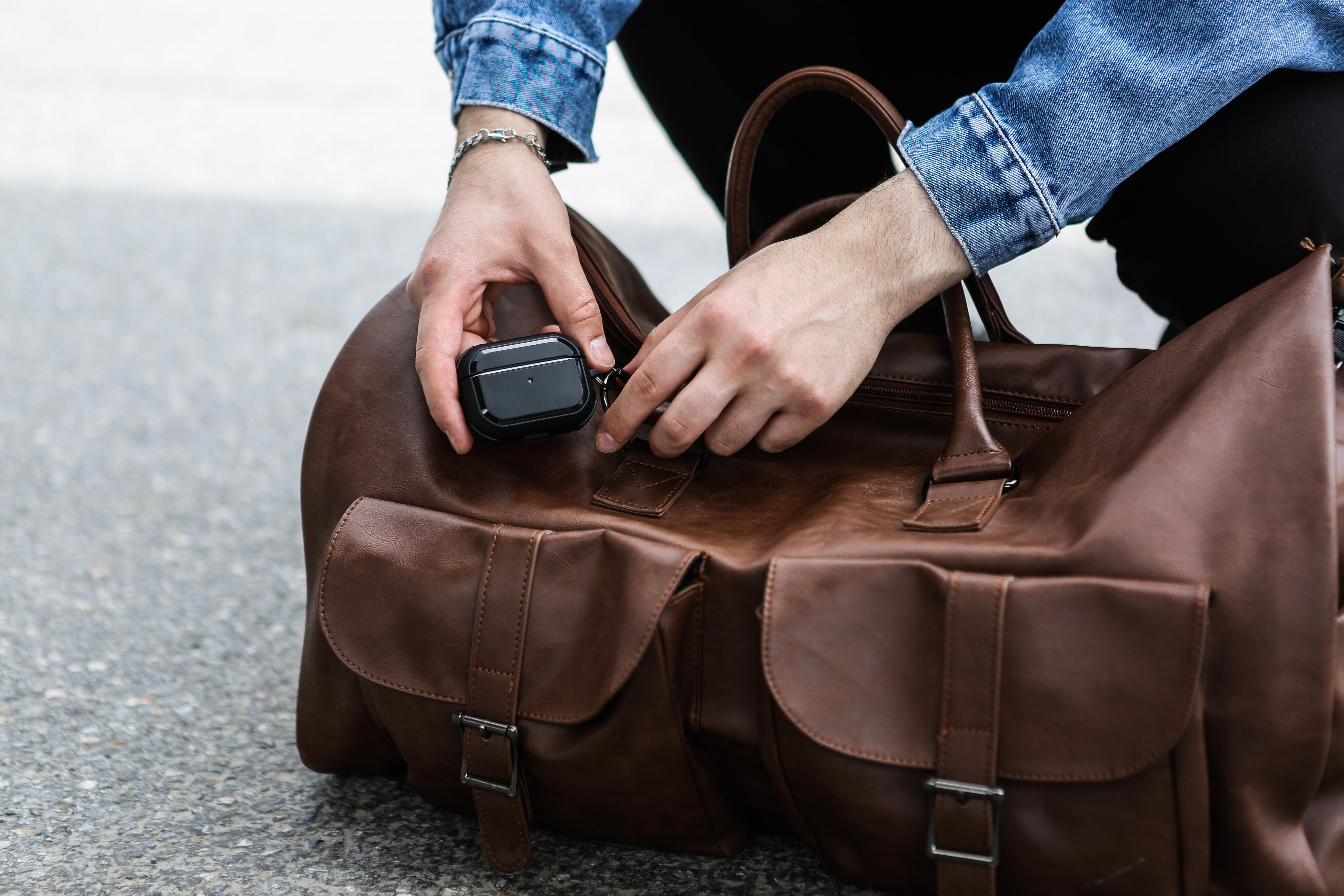 Person steckt Airpods Pro-Hülle in eine braune Leder-Duffel-Tasche. Die Tasche hat Taschen und einen Reißverschluss.