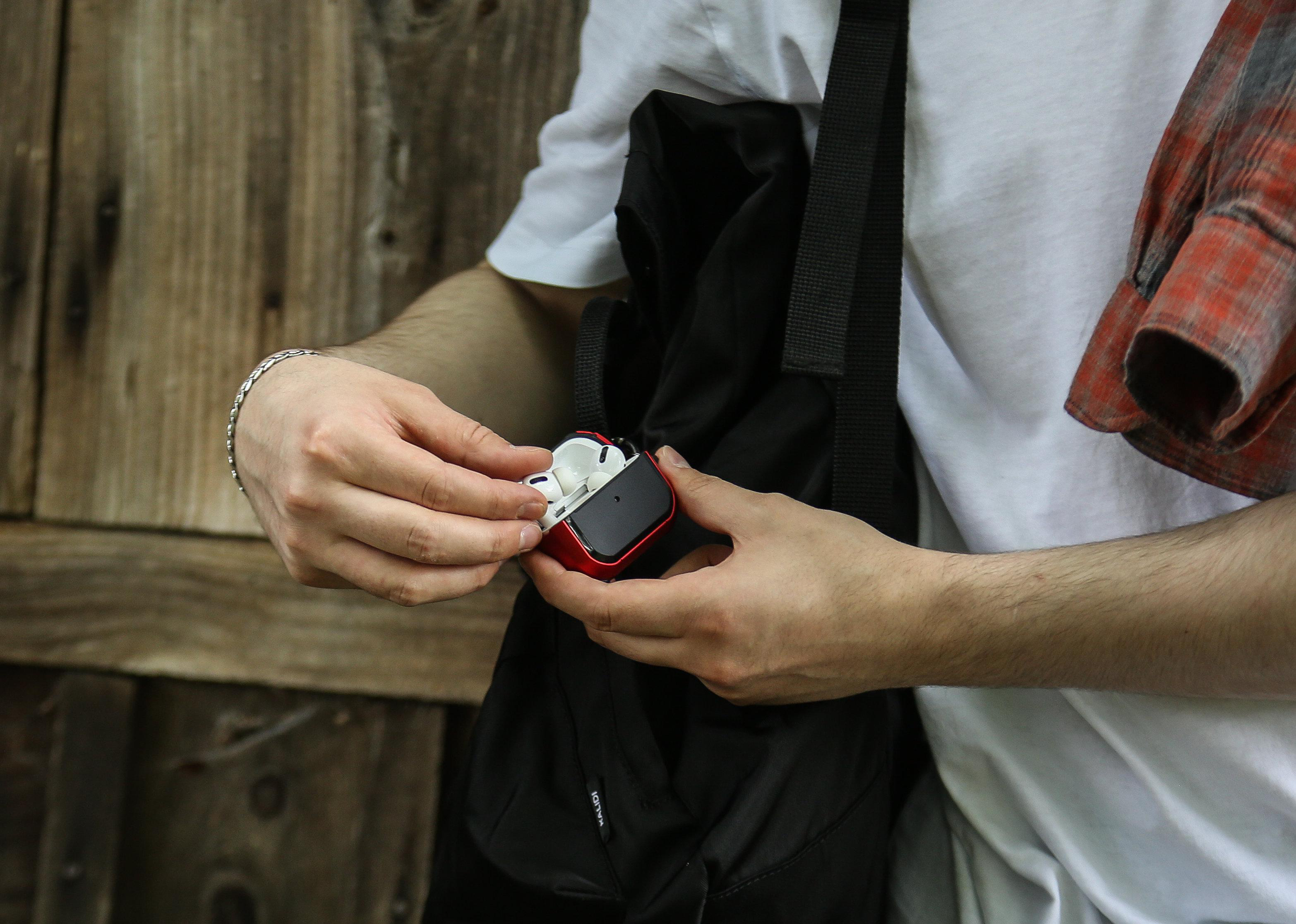 Hände einer Person, die ein rotes AirPods-Etui öffnen, neben einem schwarzen Rucksack und einem karierten Hemd.