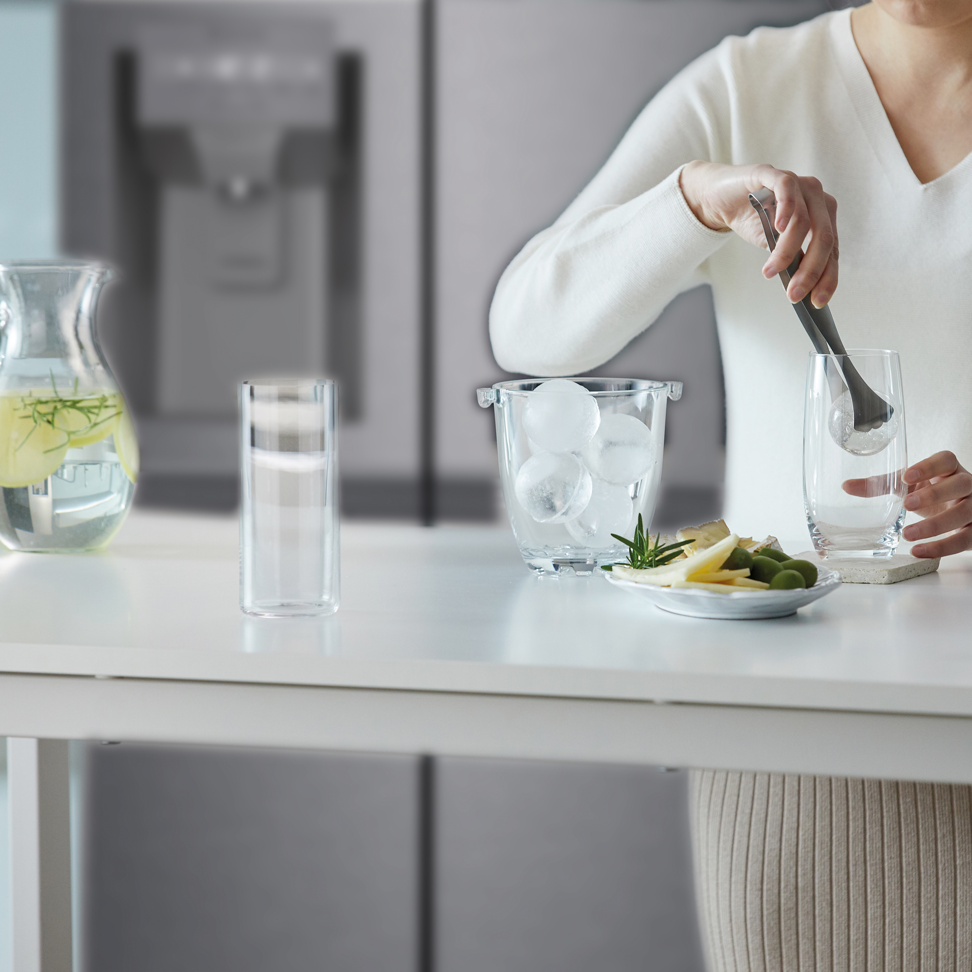 Persona usando pinzas para hielo para colocar hielo en un vaso sobre una encimera blanca con comida.