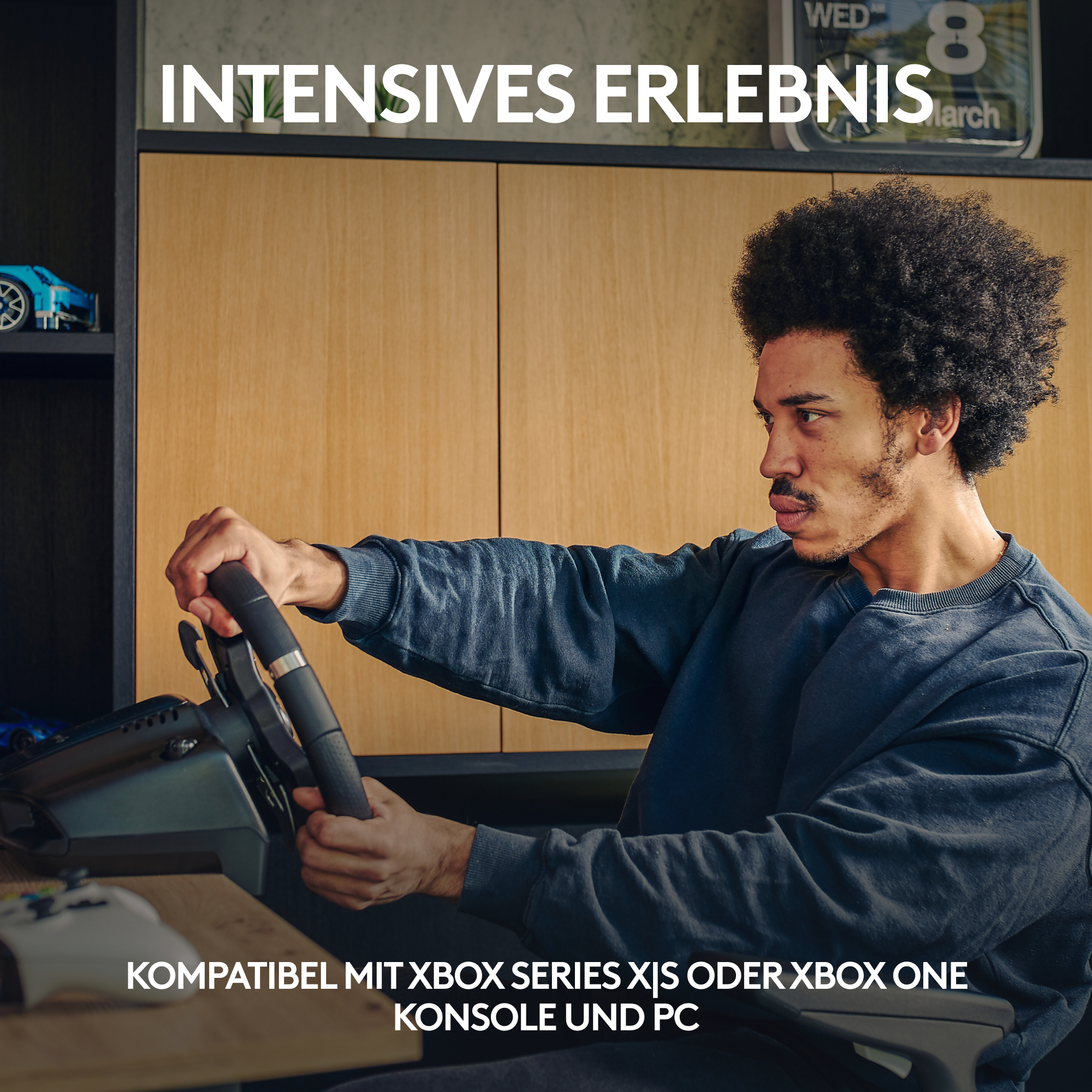 Ein Mann mit Afro-Frisur hält ein Gaming-Lenkrad vor einem Holzschrank und Textüberlagerung.