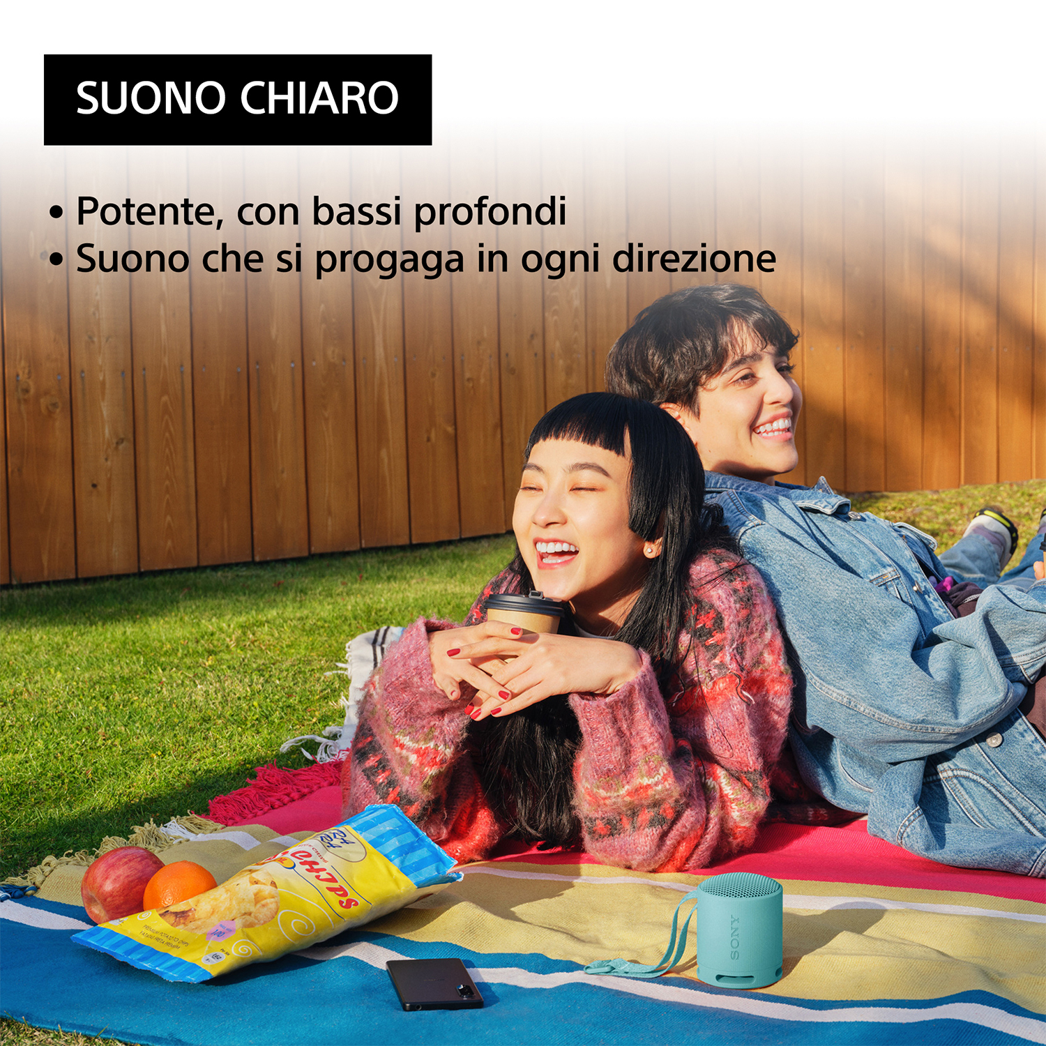 Due persone sorridenti, sedute su una coperta, con snack e un altoparlante turchese.