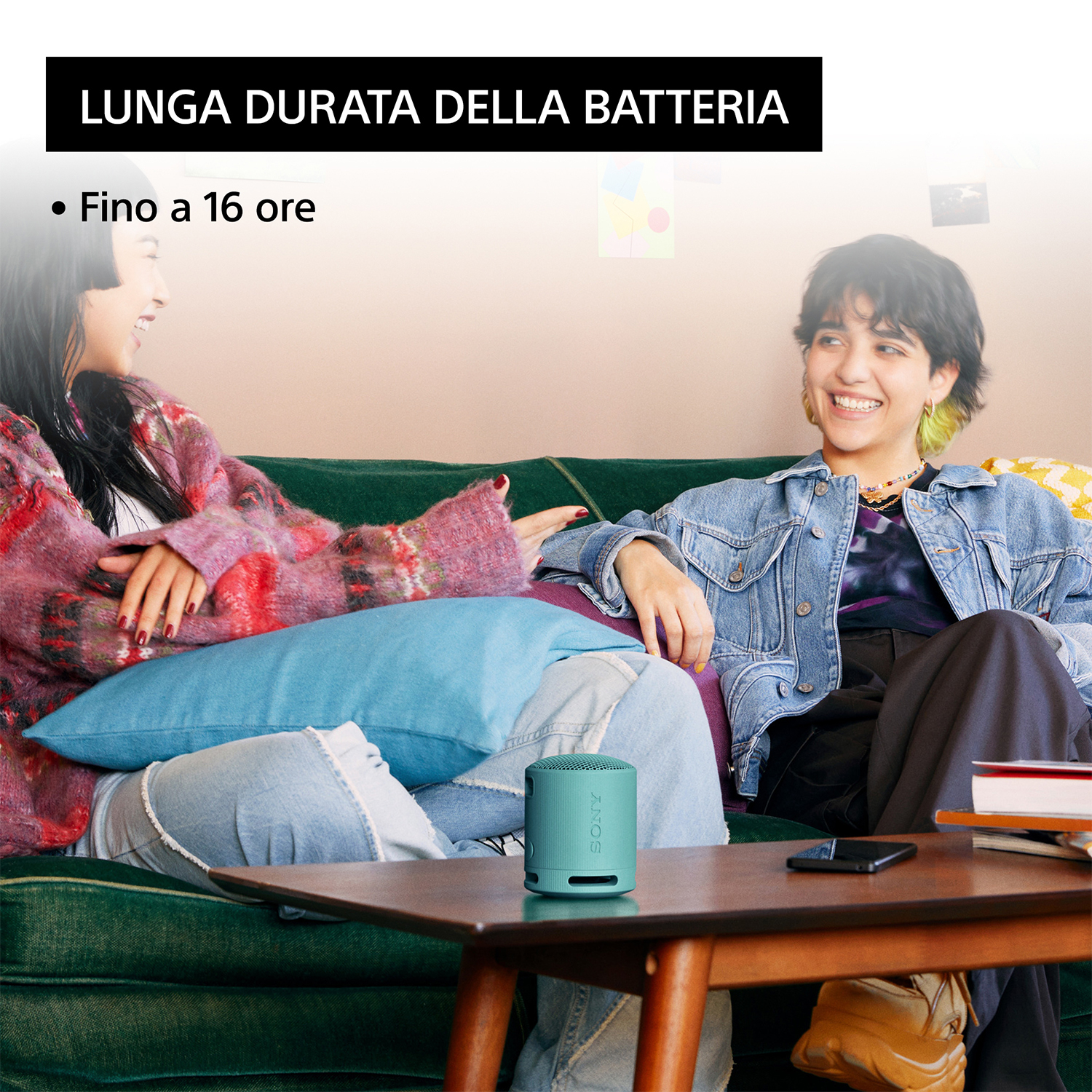 Due persone sedute su un divano verde, sorridenti, con un altoparlante blu davanti a loro.