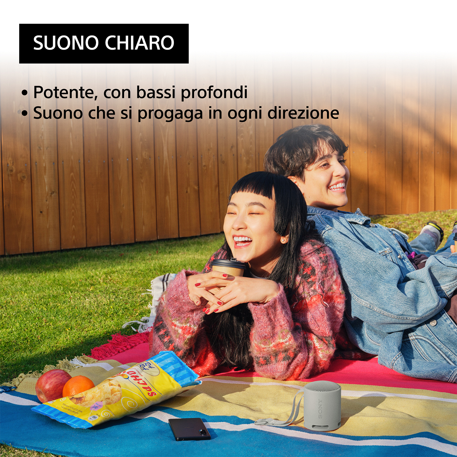 Due persone ridono, un piccolo altoparlante, un sacchetto di patatine e un telefono su una coperta.