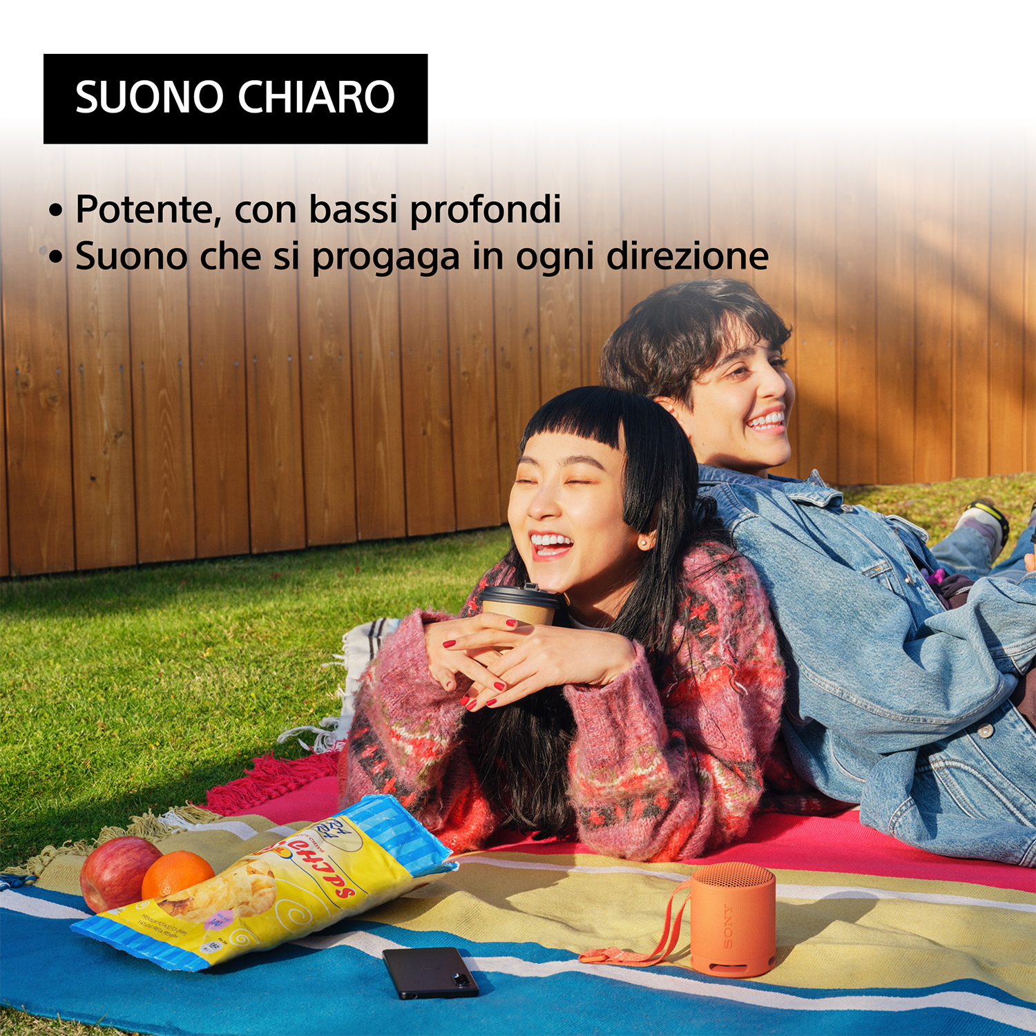 Due persone su una coperta da picnic. Una tiene una tazza di caffè, l'altra sorride. Snack e altoparlante nelle vicinanze.